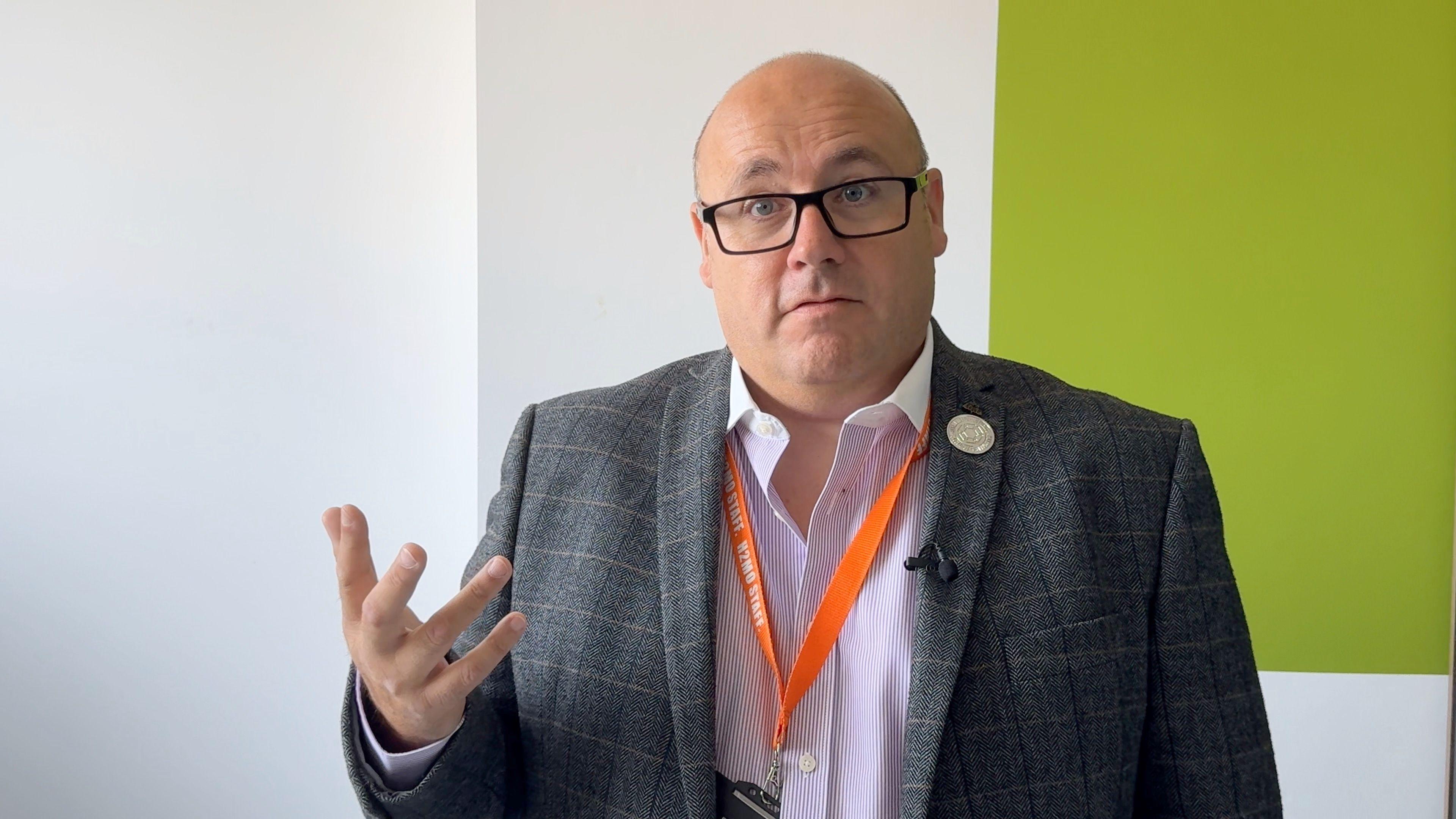 A bald man with dark glasses stands in front of a green and white background. He wears a checked suit, pink shirt and has an orange lanyard around his neck.