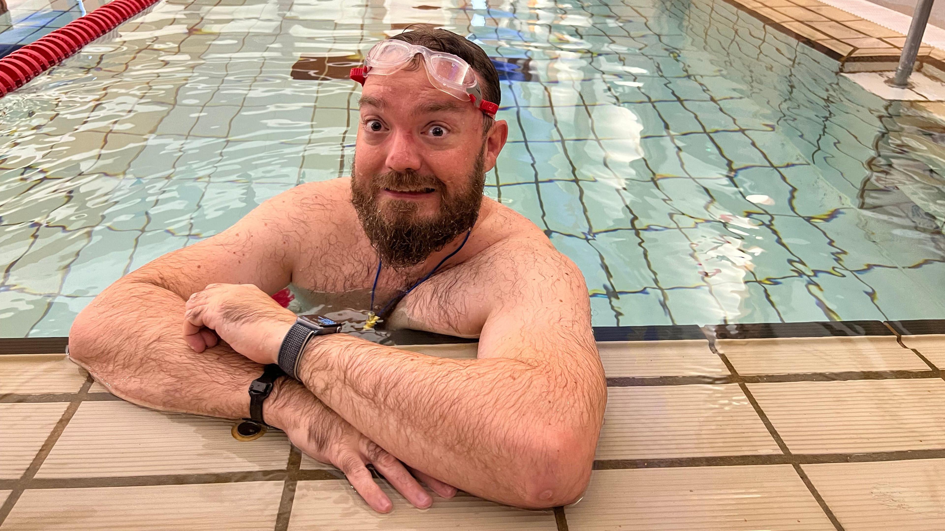 A man in a swimming pool resting his arms on the edge, wearing a pair of red goggles on his forehead and pulling a face at the camera.