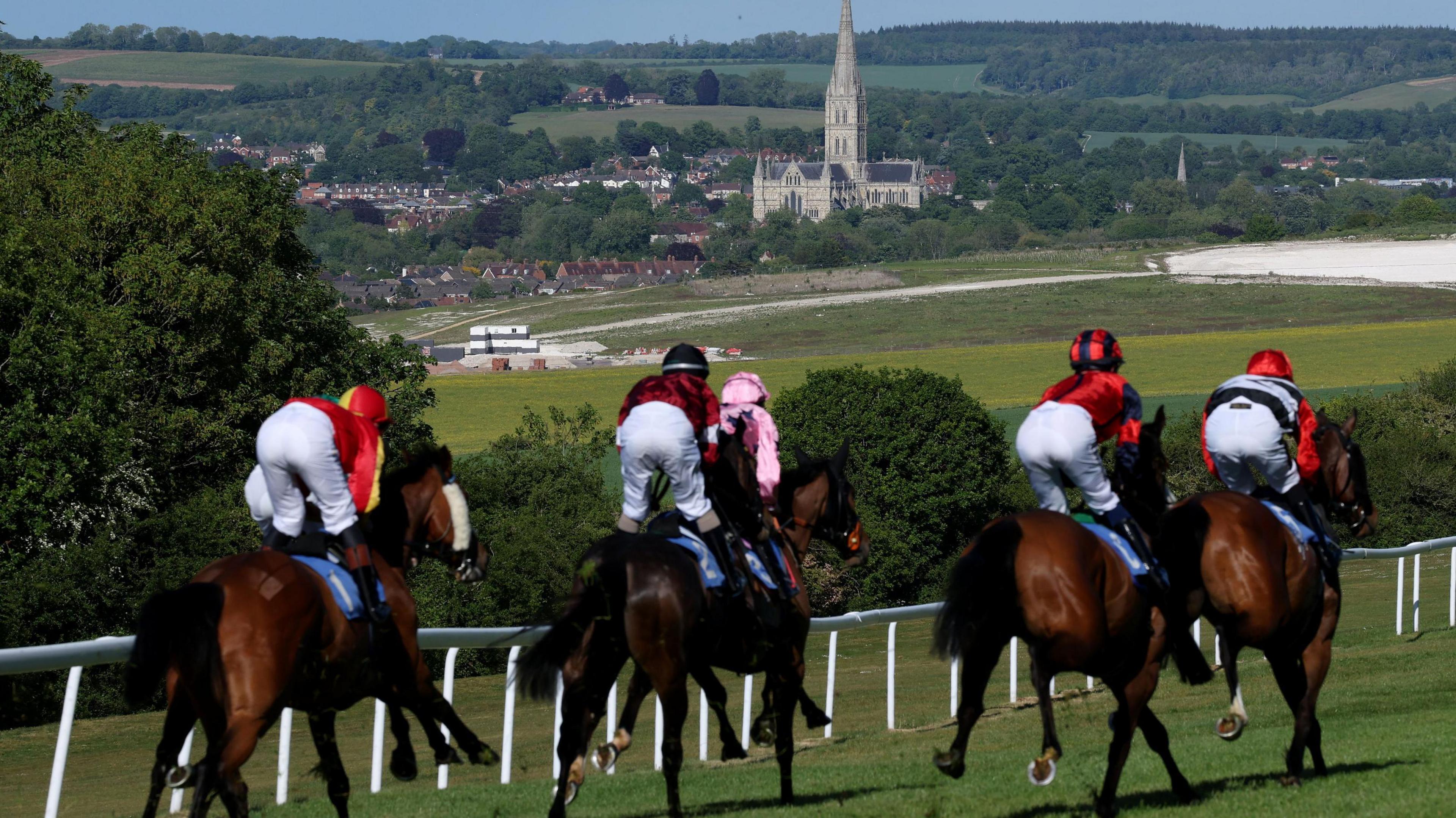 Six horse riders head down the back straight against the backdrop of Salisbury Cathedral. They are wearing riding gear of pink and red and on brown horses.