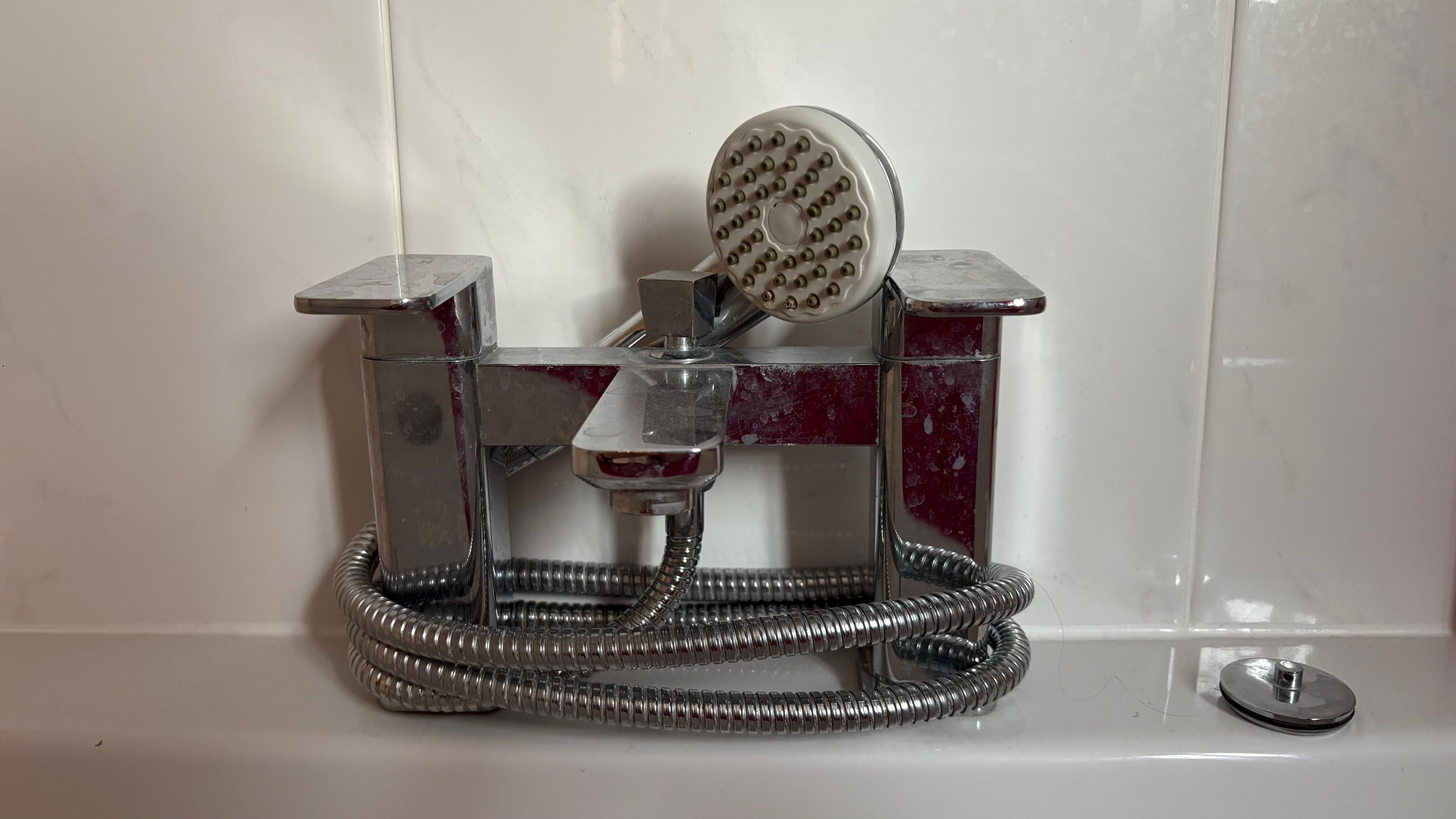 A modern mixer bath tap showing rectangular handles and a central spout with adjuster at the back. There is also a shower head on the end of a flexible gooseneck which is wound around the taps. There are bathroom tiles behind.