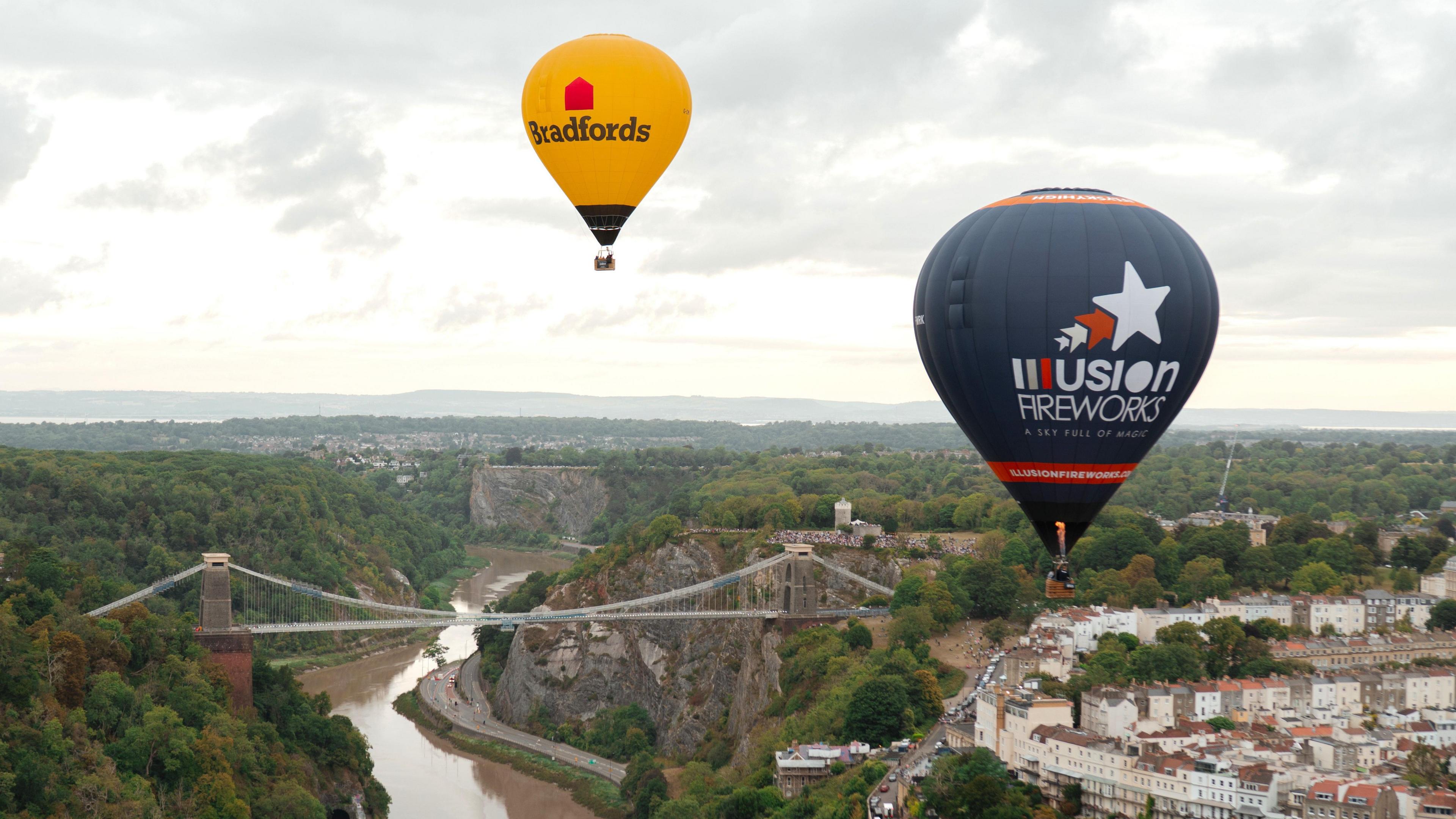 Two hot air balloons, one bright yellow and one dark purple, are in the air close to the Clifton Suspension Bridge after a mass ascent at the Bristol International Balloon Fiesta. The River Avon is visible under the bridge, along with housing in Clifton.