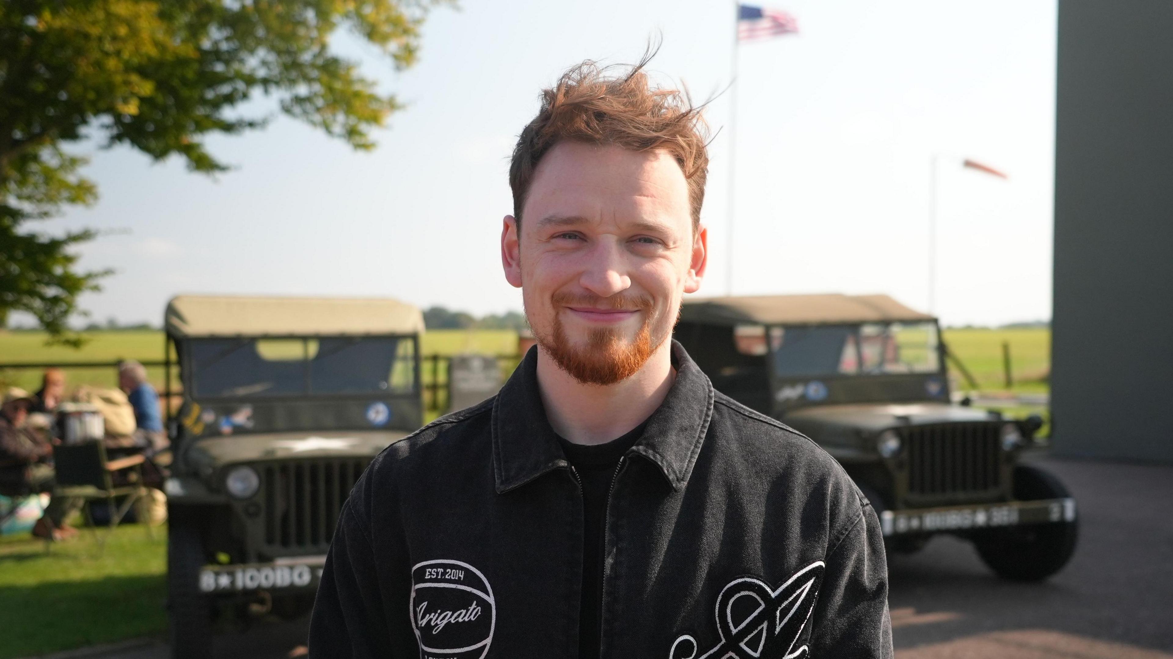 Louis Greatorex wearing a black shirt and standing in front of two military vehicles and an American flag in the distance