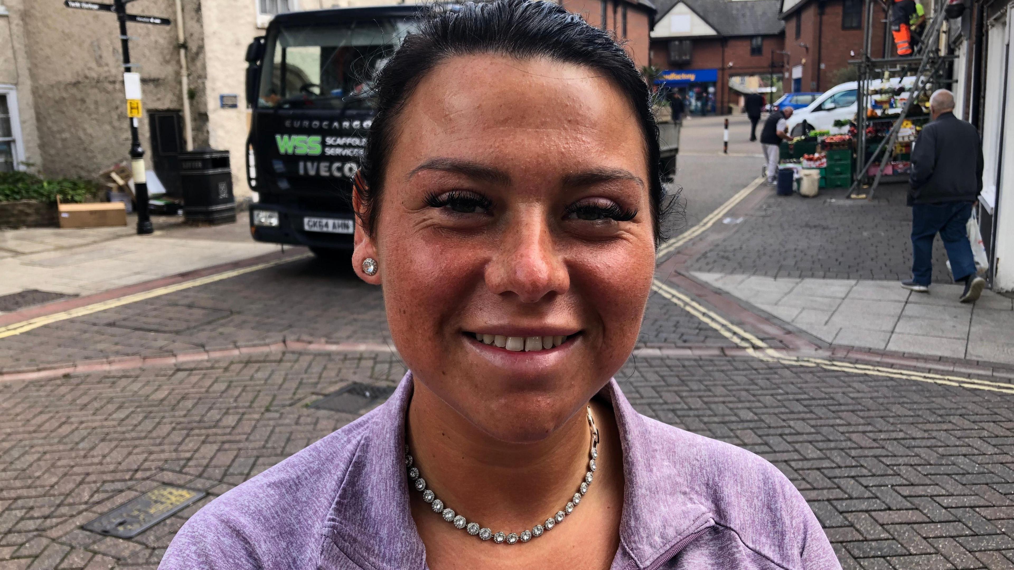 A smiling Abbie Chambers looks directly at the camera in a purple top and silver necklace. She is photographed in a street. 