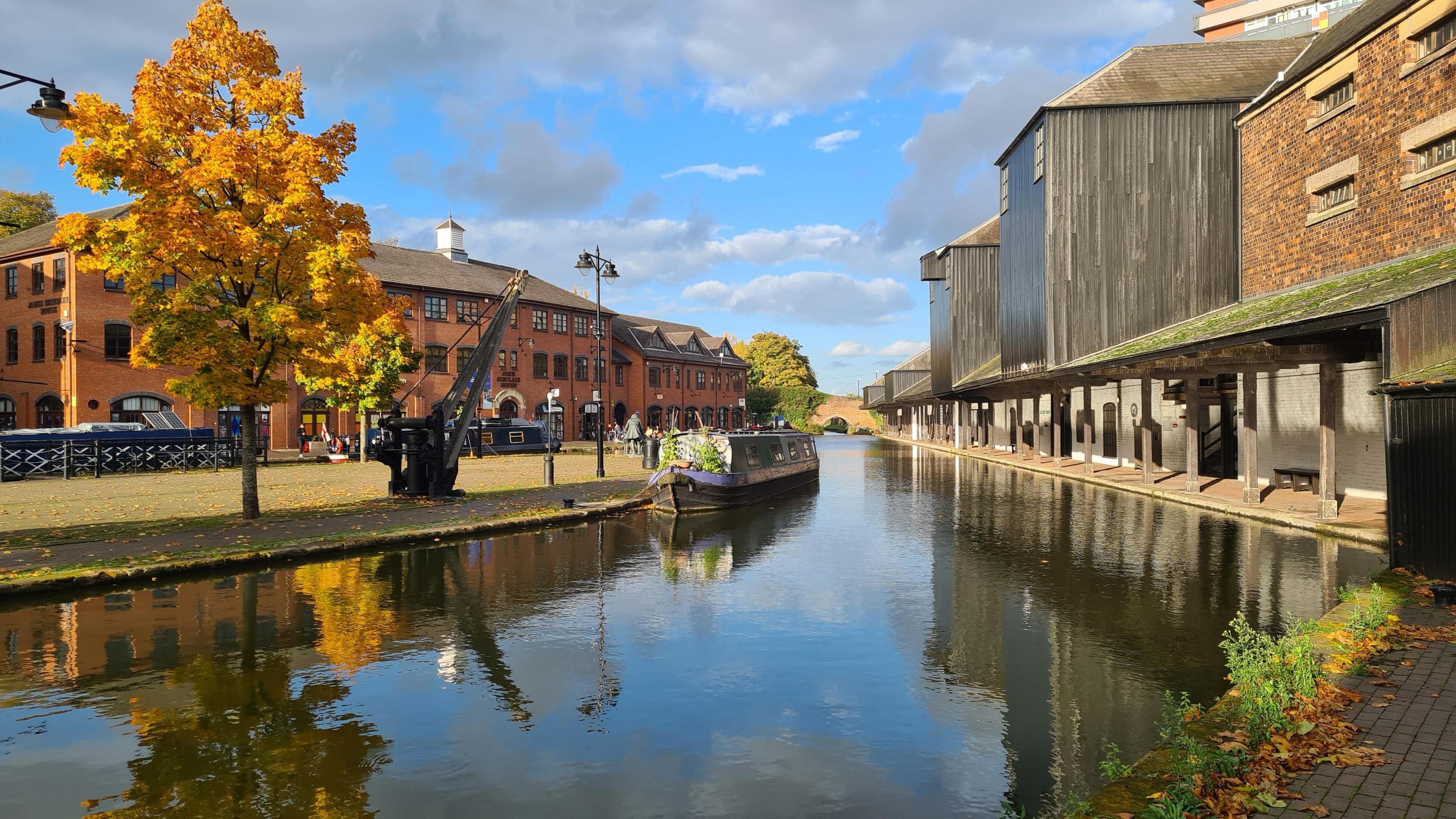 A canal basin with restored and rebuilt industrial buildings around it and a boat moored in the centre of the picture. A tree with yellowing leaves overlooks the water beneath a blue sky punctuated by grey clouds. 