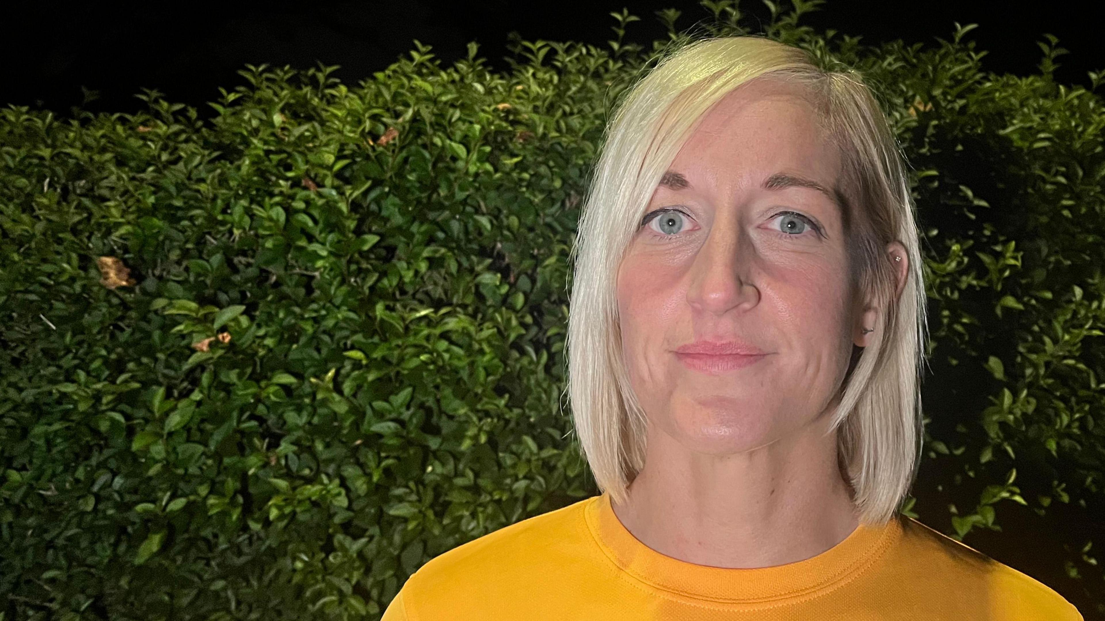 Runner Gemma Stephenson is wearing an amber-coloured T-shirt. She has blond shoulder-length hair. She is standing in front of a green shrub under a night sky.