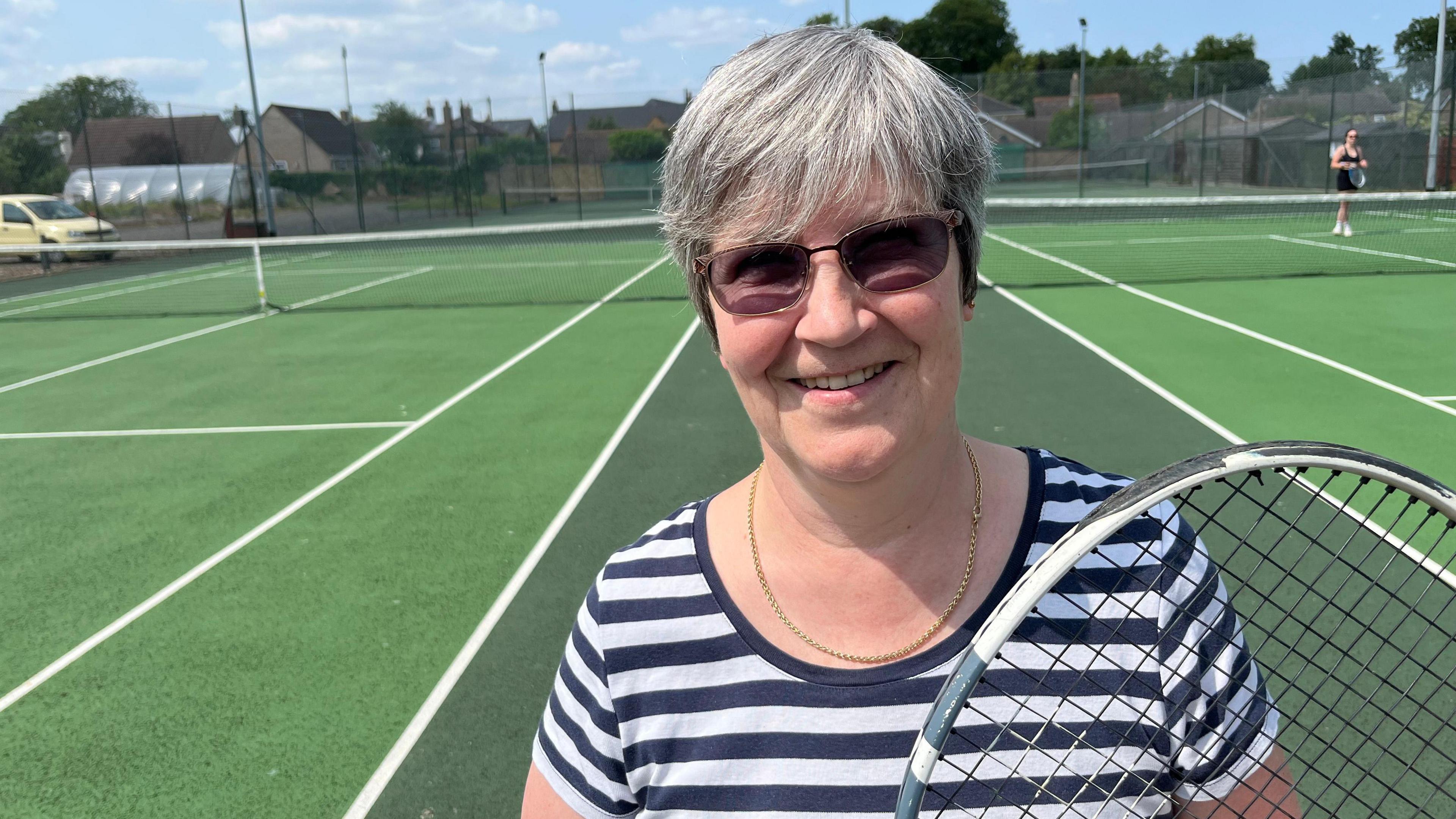 Anne Appleby, 60, has light brown hair and is wearing a black and white hooped round collared T-shirt, with a gold chain visible around her neck. She has dark glasses on and is holding a tennis racquet. She is standing on a tennis court.