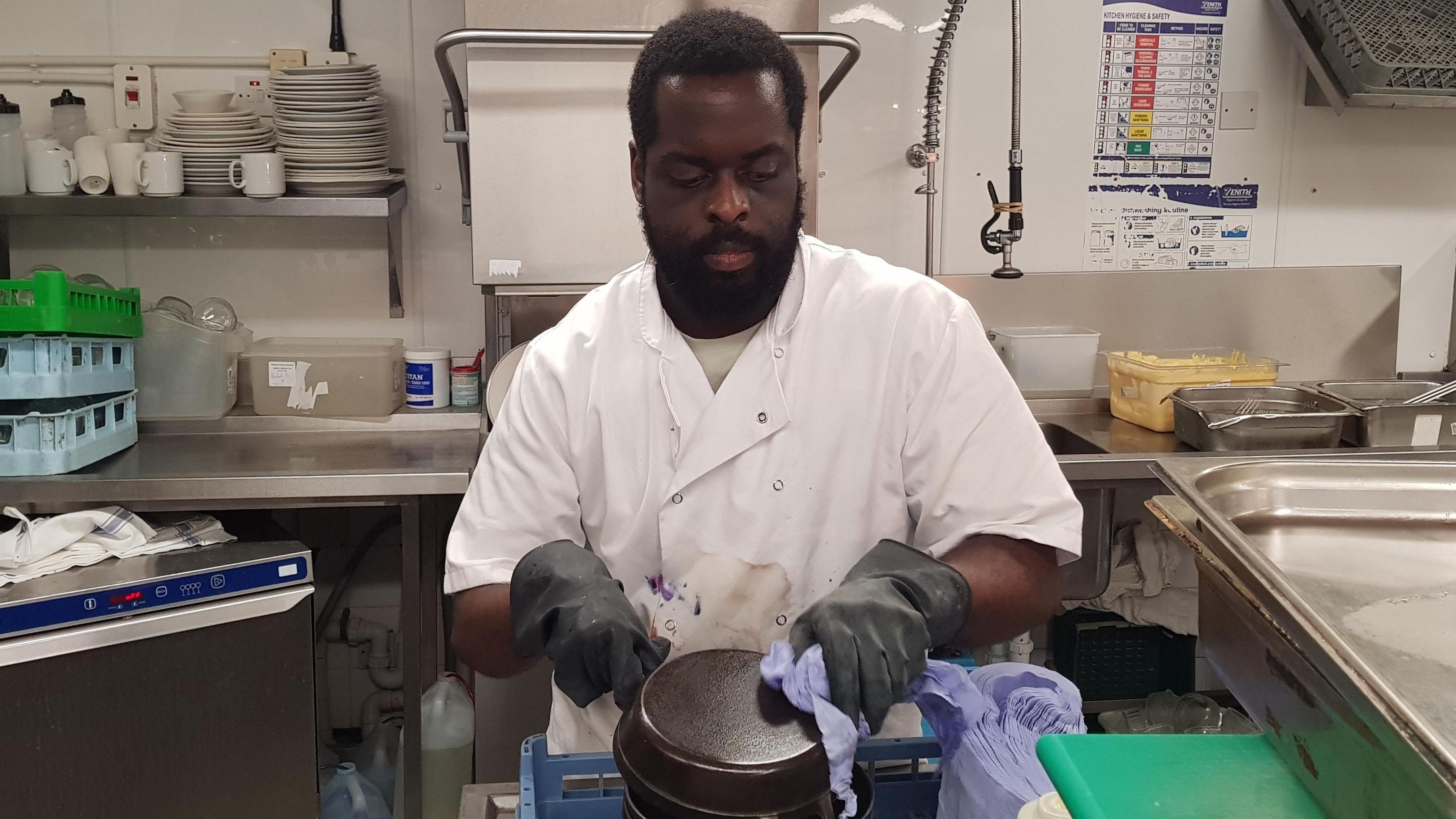 Jomi Denedo working in a kitchen, he is wearing a white chefs suit, and black gloves, he is cleaning a pan. Kitchen items are around him including paper towels, a sink and plates and cups on a shelf to the left. He is looking down, and has a beard. 