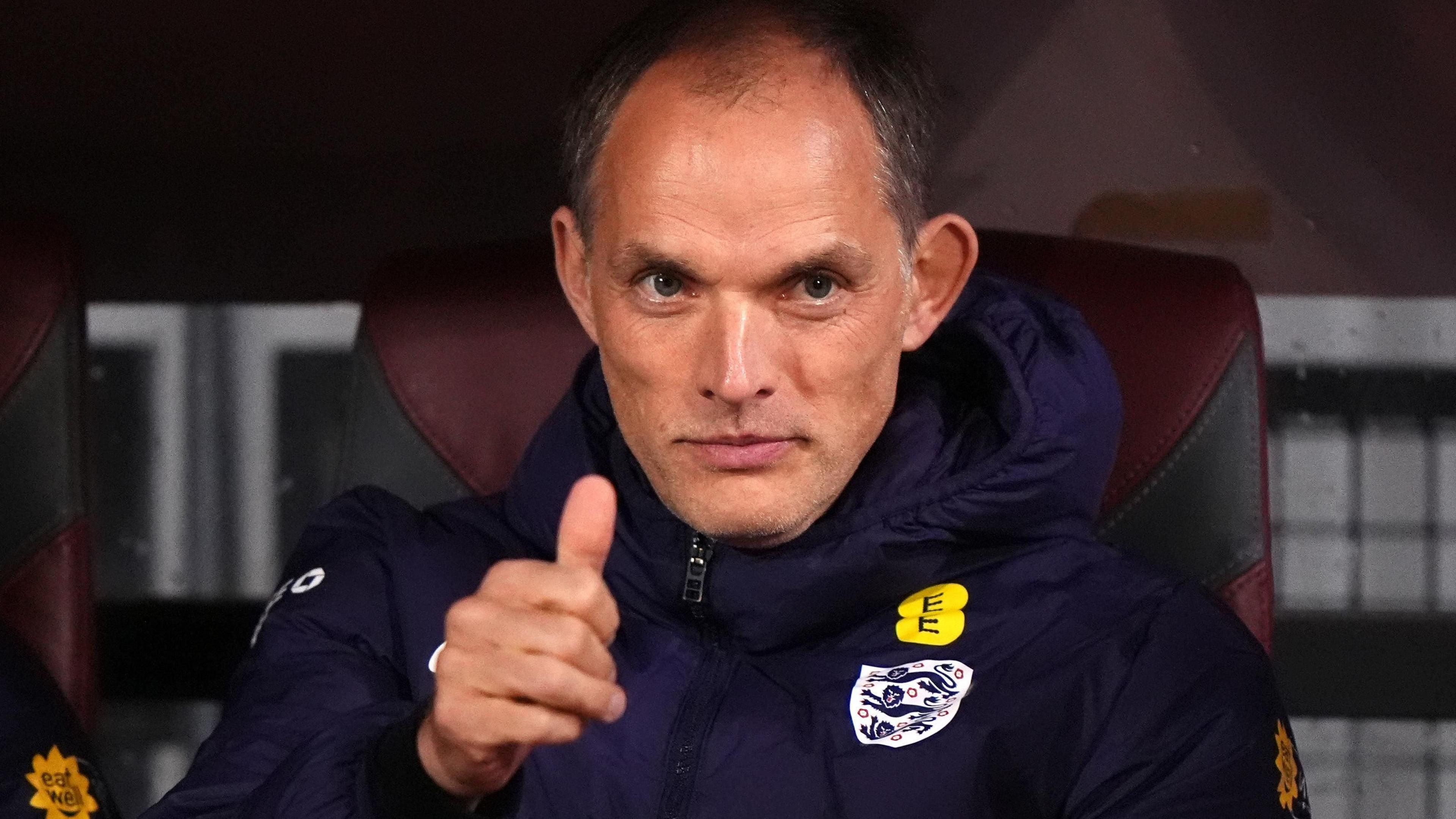 England Manger Thomas Tuchel wears a navy puffa jacket with the three lions logo, offering a thumbs up to the camera.