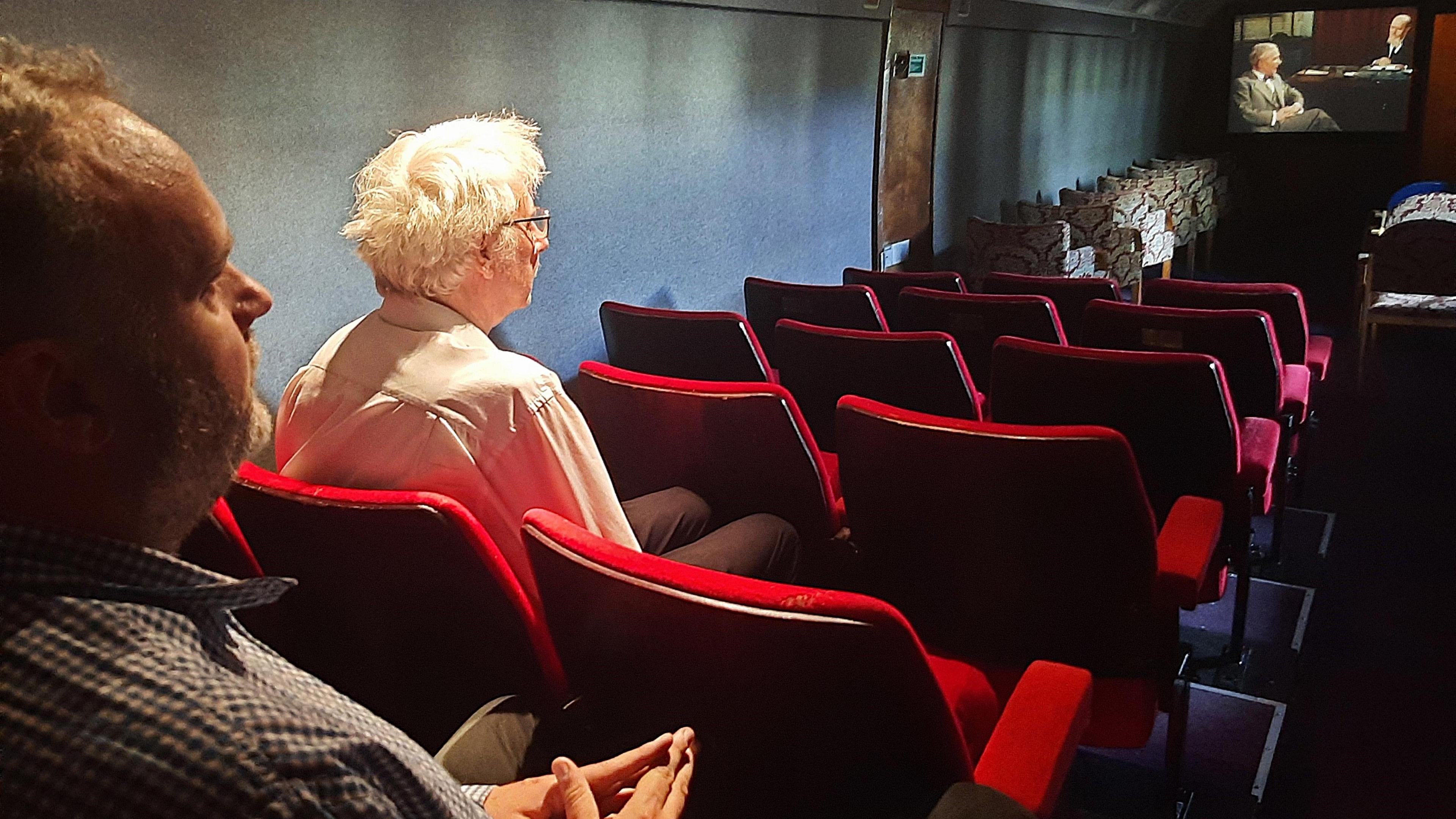 A view slightly from behind of the now restored carriage, with vintage red cinema seats and a projector screen in the distance with a film being shown. Rob and Steve Foxon are at the back, their side profiles can be seen from this angle