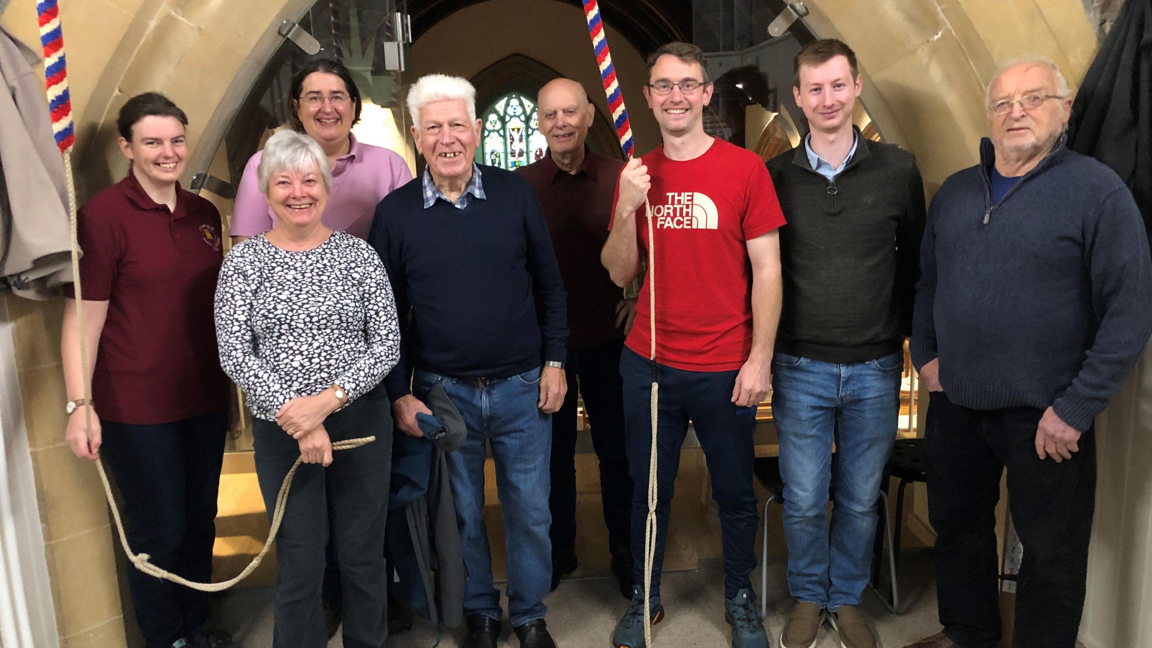 A group of people stand in a church belfry. They hold the ropes of the church bells. An older man with white hair, a dark sweater and blue jeans stands fourth from the left. A stained glass window is visible in the background.