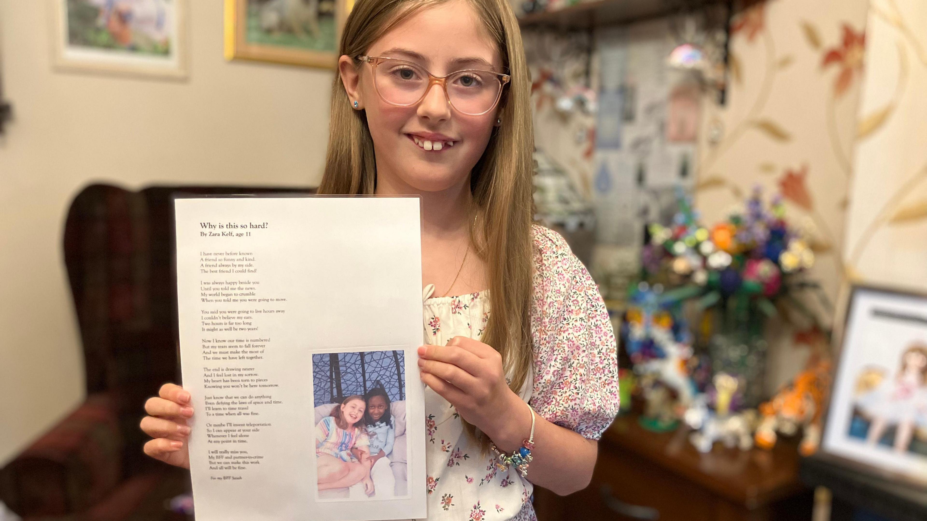 11 year old Zara pictured in a living room holds up a typed, laminated copy of a poem. There is an image of two girls (her and her friend) on the sheet.