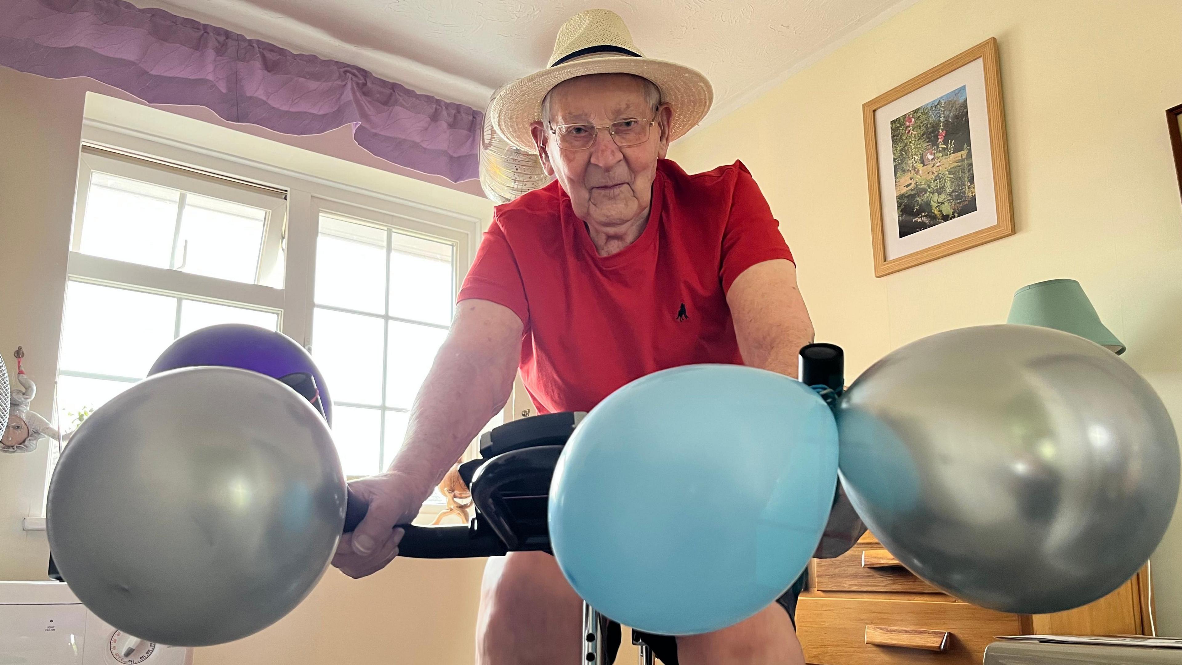Geoffrey Bolton, 100, on a exercise bike with coloured balloons tied to the handle bars, he is wearing a white fedora hat, black shorts, red top, and glasses. Behind him is a window and pictures on the walls.