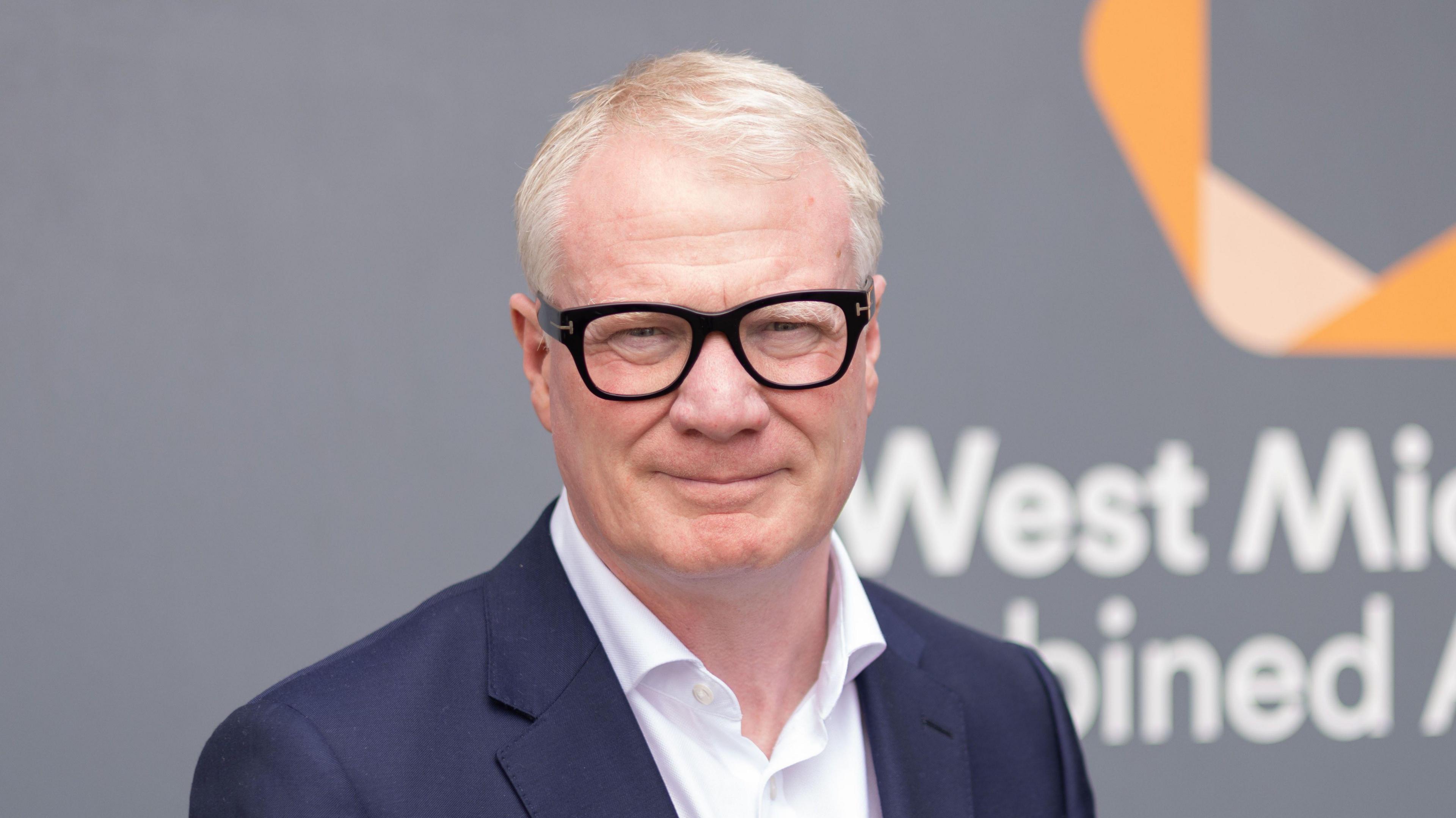 West Midlands Mayor Richard Parker smiles at the camera, in a close-up of his face and shoulders. He stands in front of a grey board which says West Midlands Combined Authority on it.