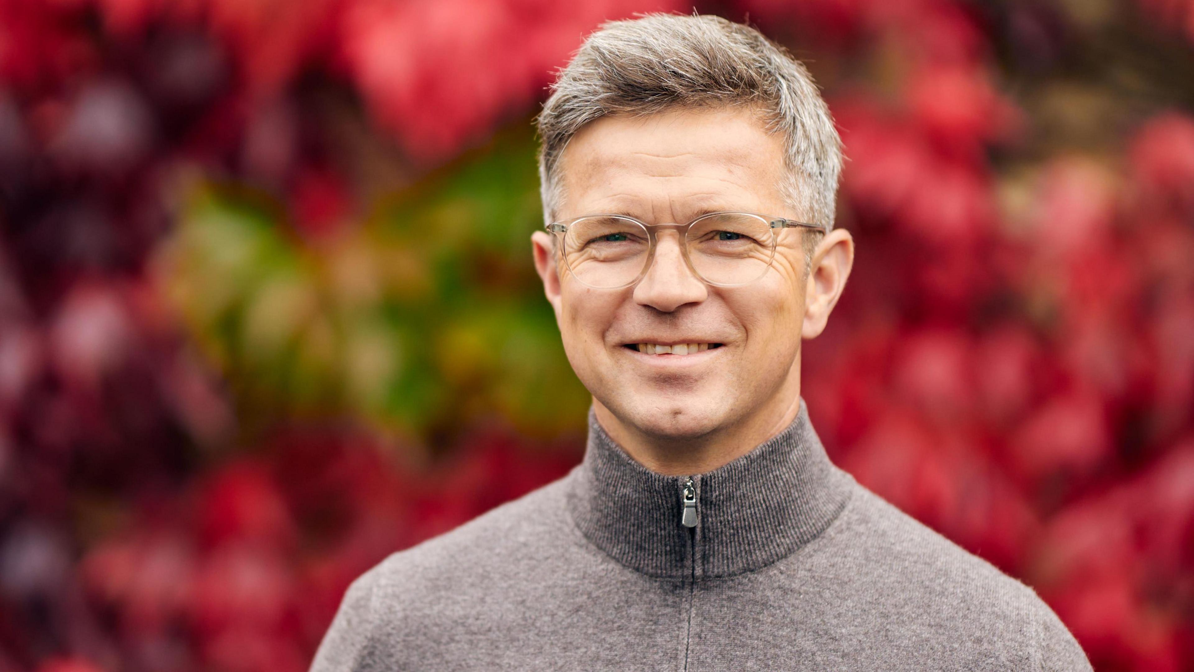 Roger Varian, wearing a grey quarter-zip sweater, smiles at the camera. He has short grey hear and glasses. A red-coloured hedge can be seen behind him.