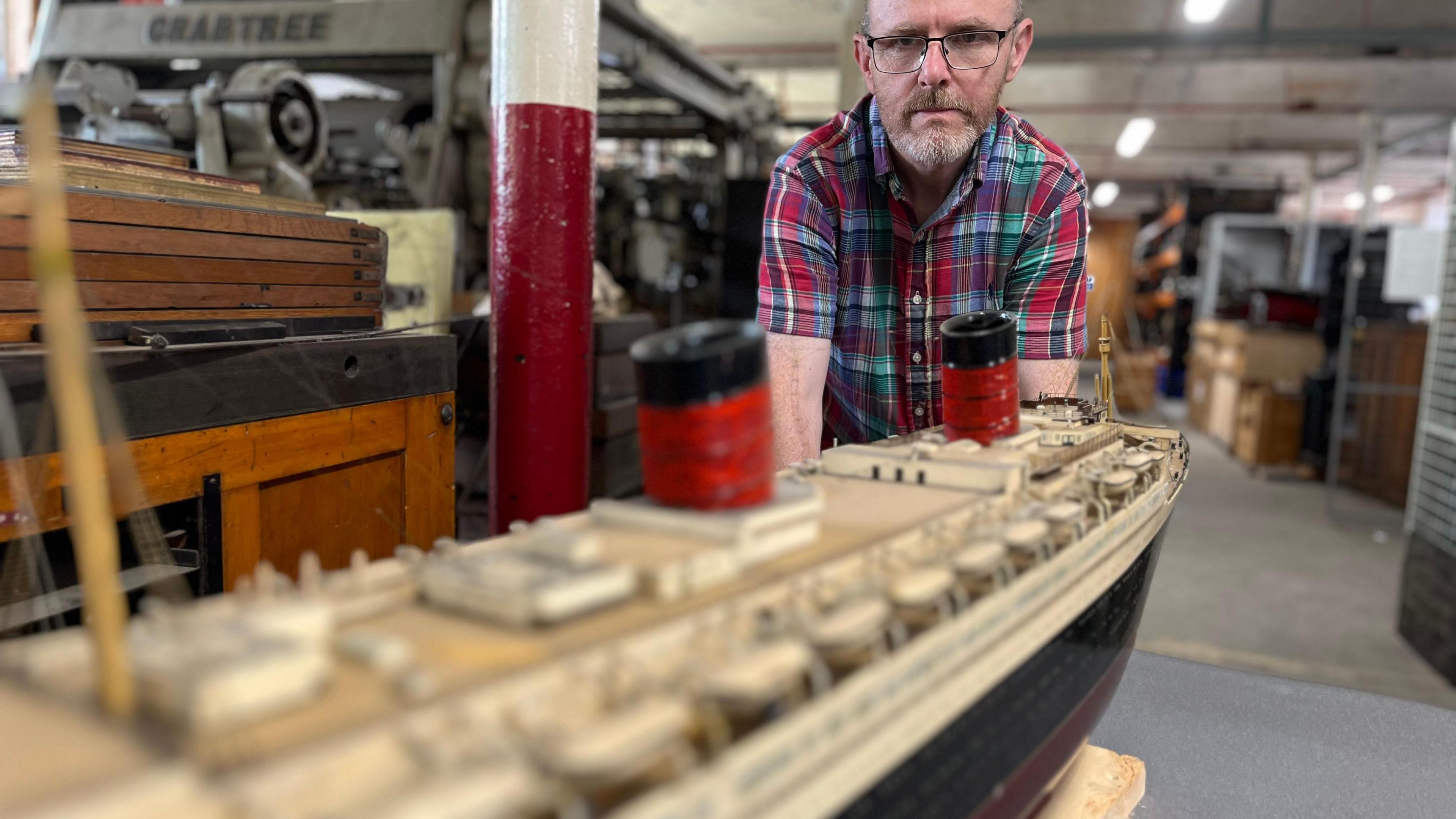 A man peers at a model of the RMS Queen Elizabeth. He is wearing a checked shirt and glasses. 