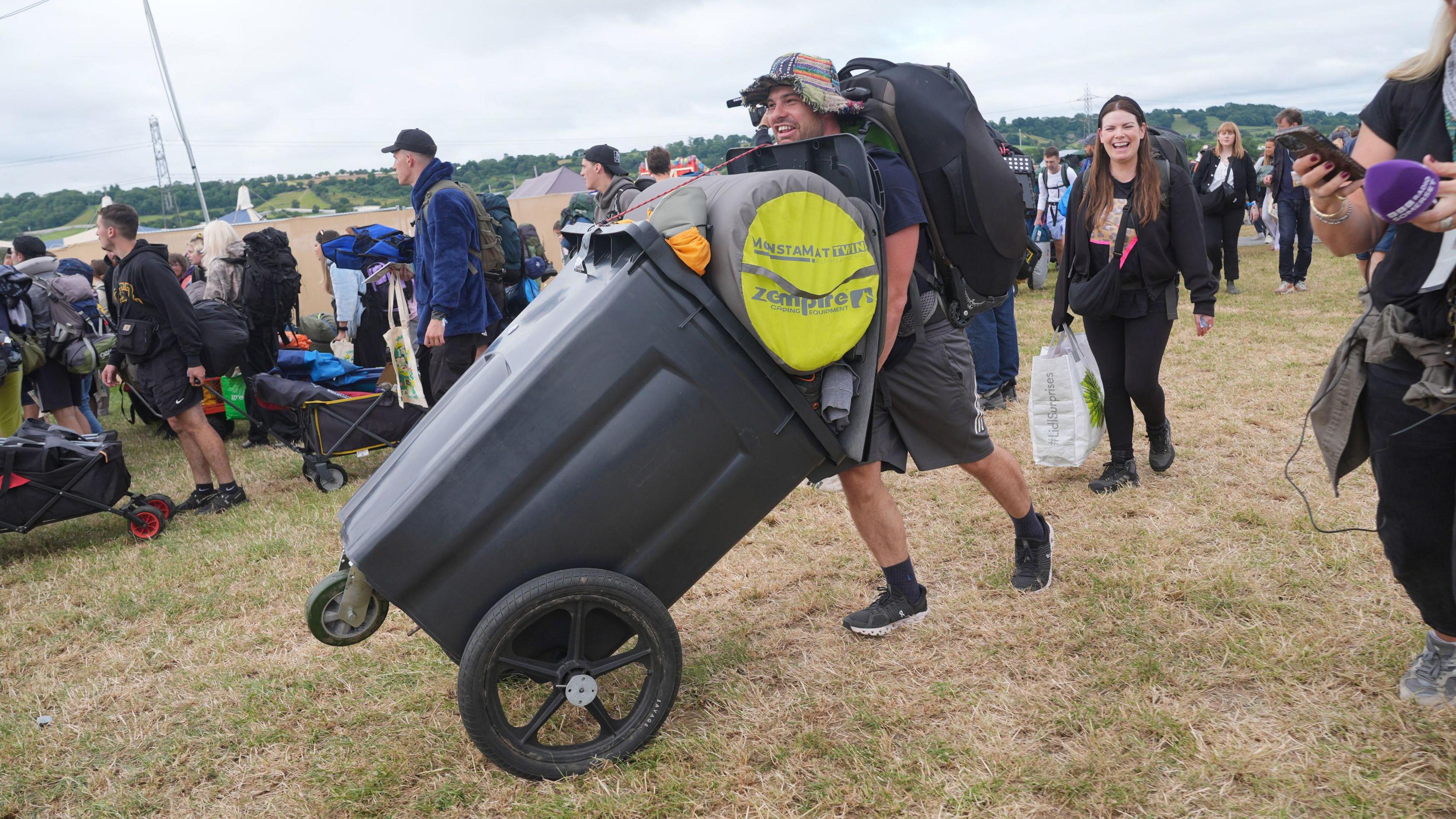 Man pushes modified wheelie bin across grass with bags and sleeping equipment in
