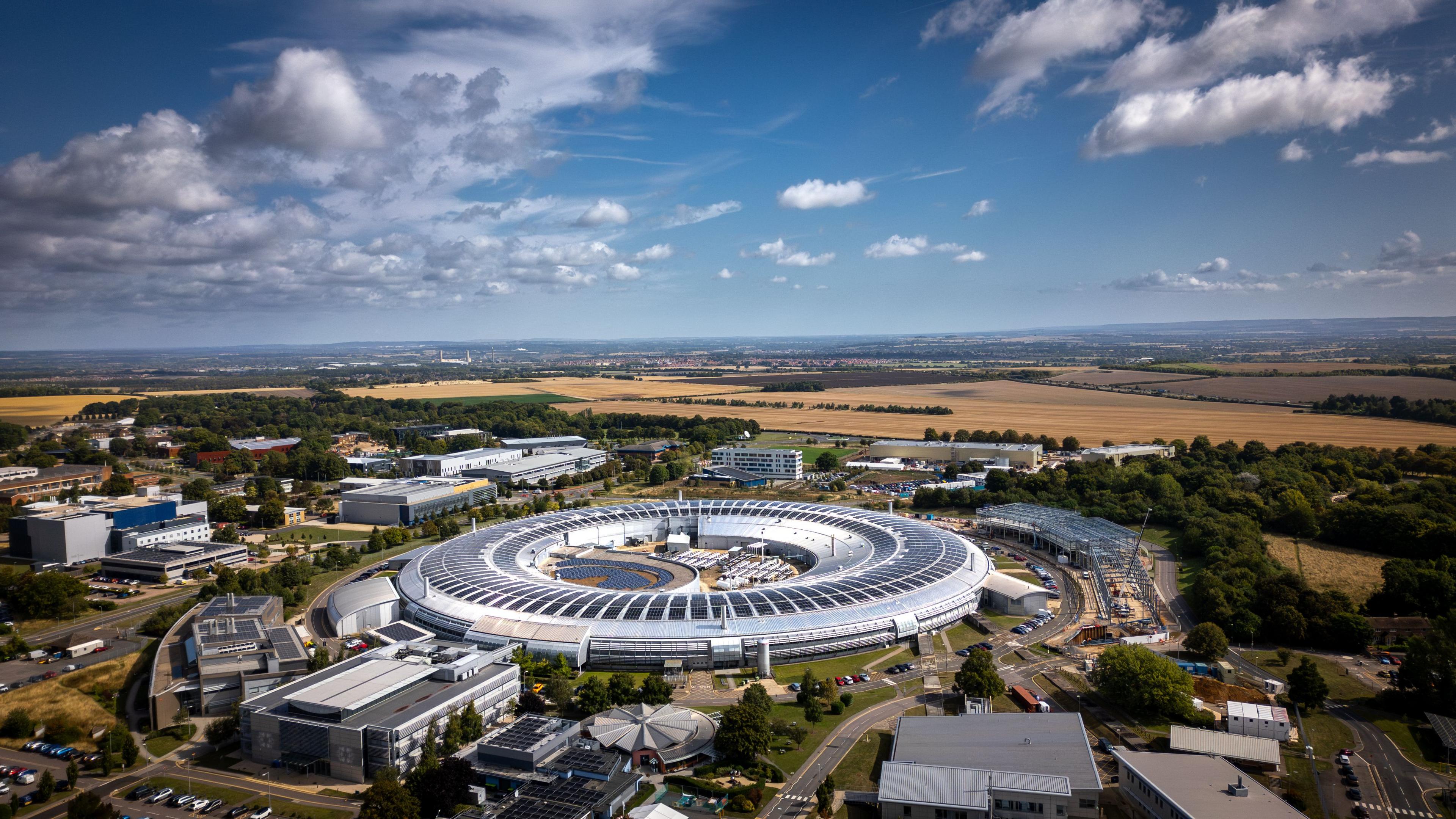 Arial image of Diamond Light Source in Oxfordshire. The building is a large, silver donut-shaped ring which houses a particle accelerator inside. The countryside can be seen surrounding the machine. 