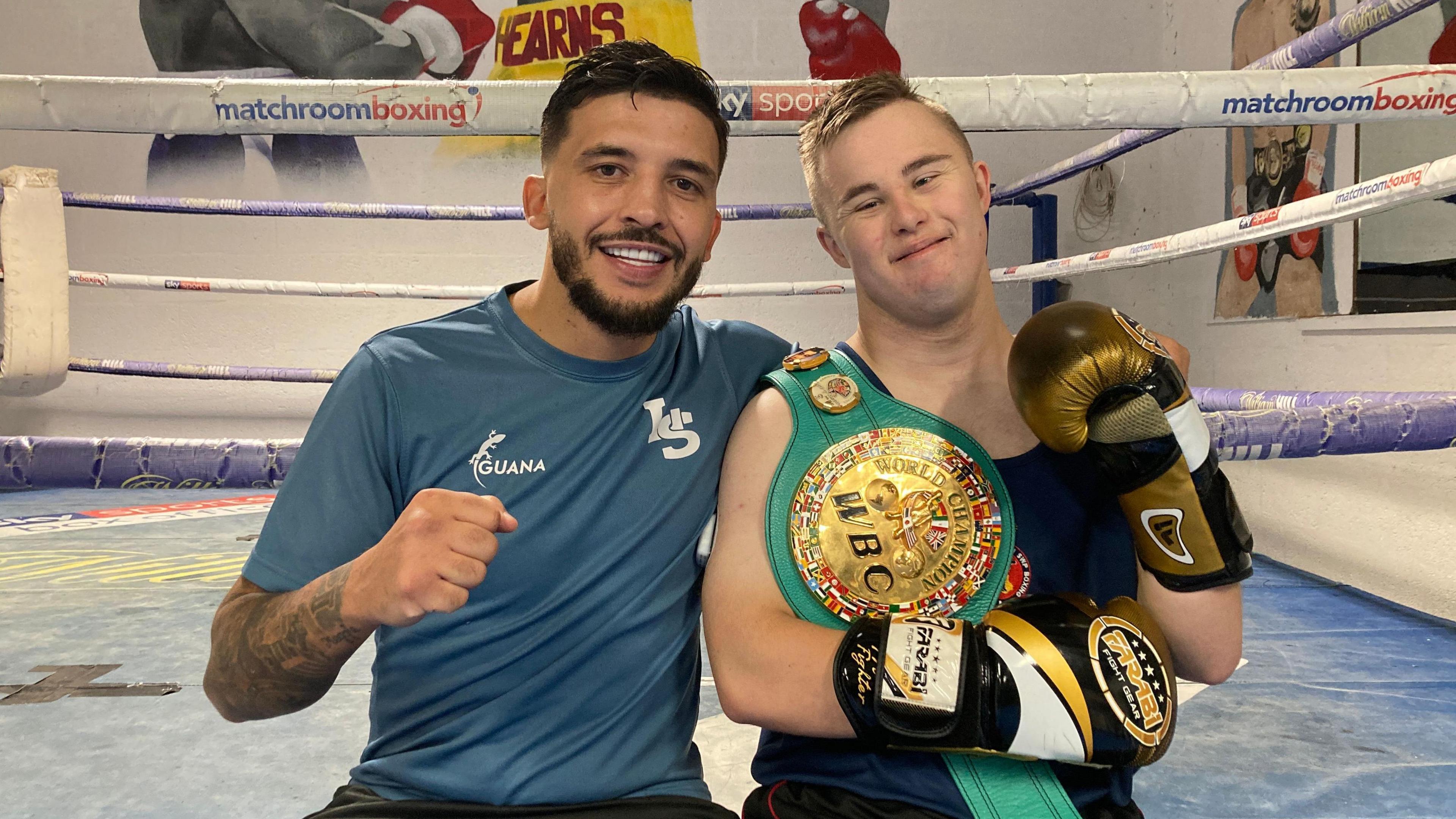 Lee Selby sat next to Jake Edwards in front of a boxing rind. Jake is wearing a winner's sash and gold boxing gloves