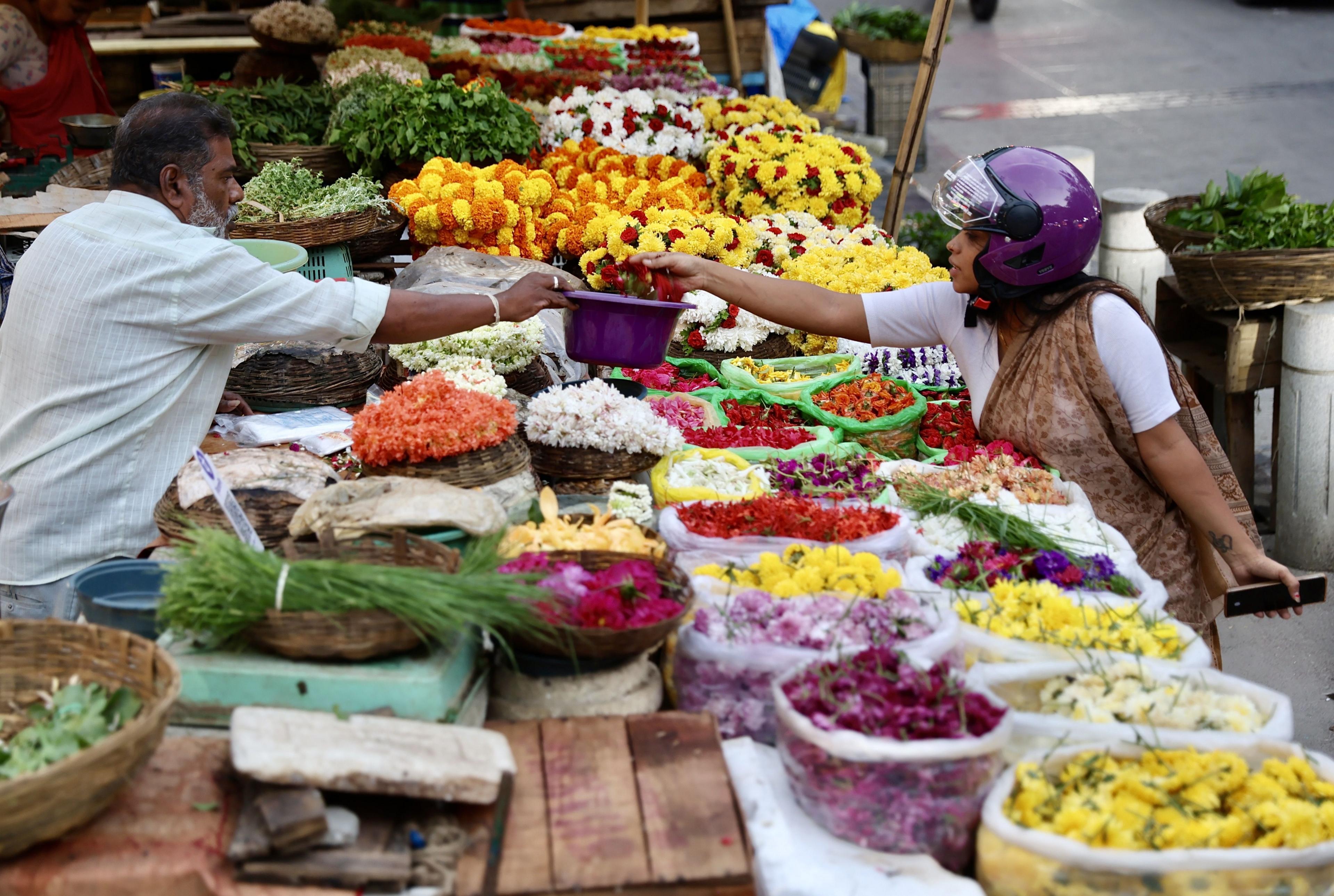 a woman wearing a motorcyle helmet reaches across a table laden with very colourful flowers and picks up some flowers from a bowl as a seller offers them to her