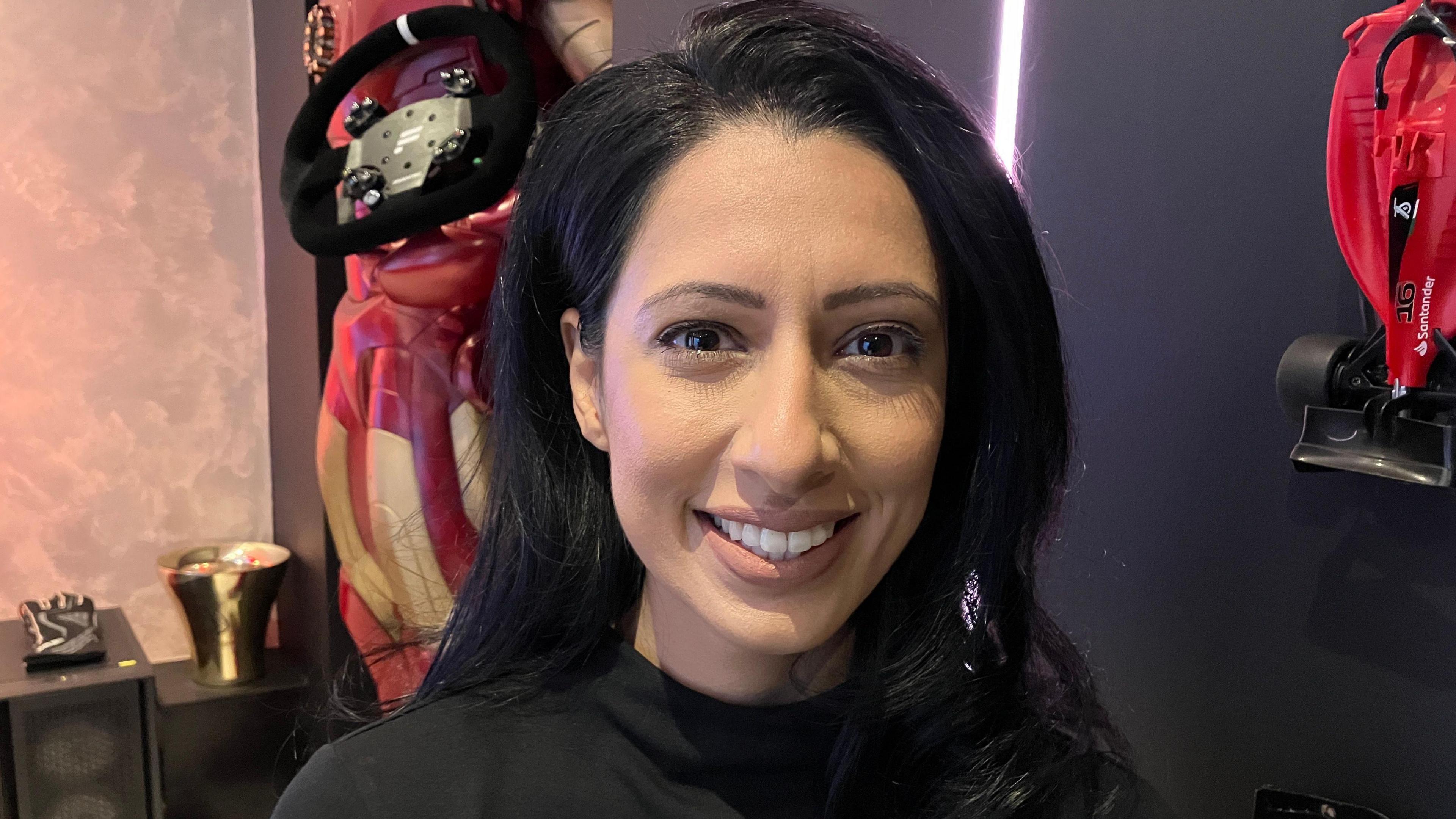 Amardeep has long black hair and is wearing a black top. She is smiling at the camera. Behind her is a small red Ferrari f1 car model hanging on the wall, next to a larger model which is also on the wall.