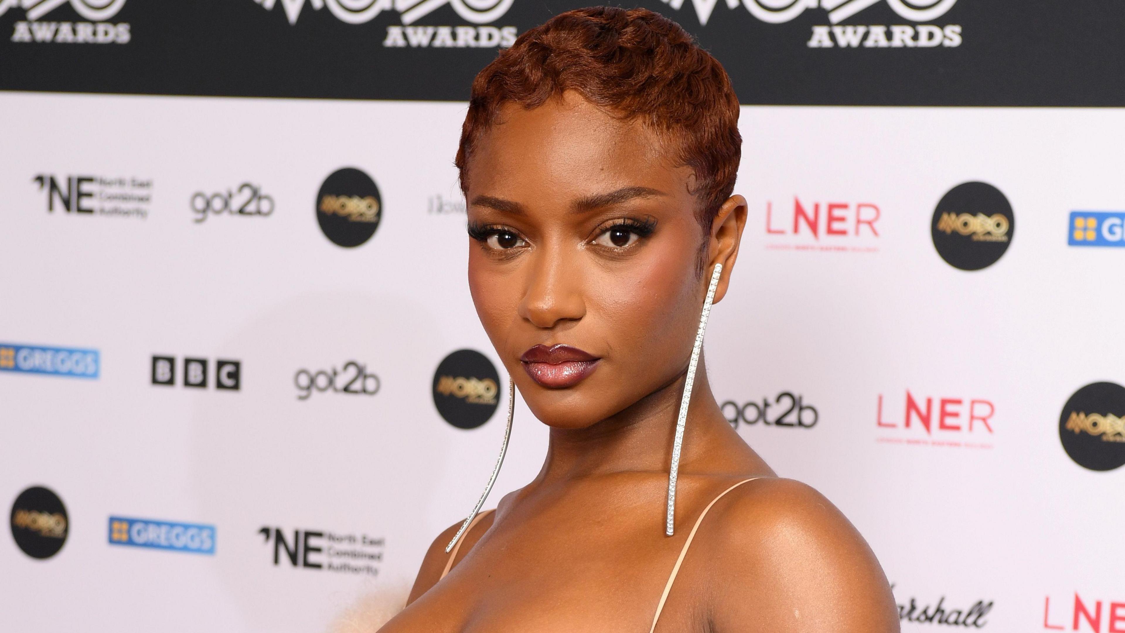 Ayra Starr at the Mobo Awards. She wears a pale pink strappy dress matched with long dangly earrings which reach her collarbone. She has short red hair and looks at the camera with a serious expression. 