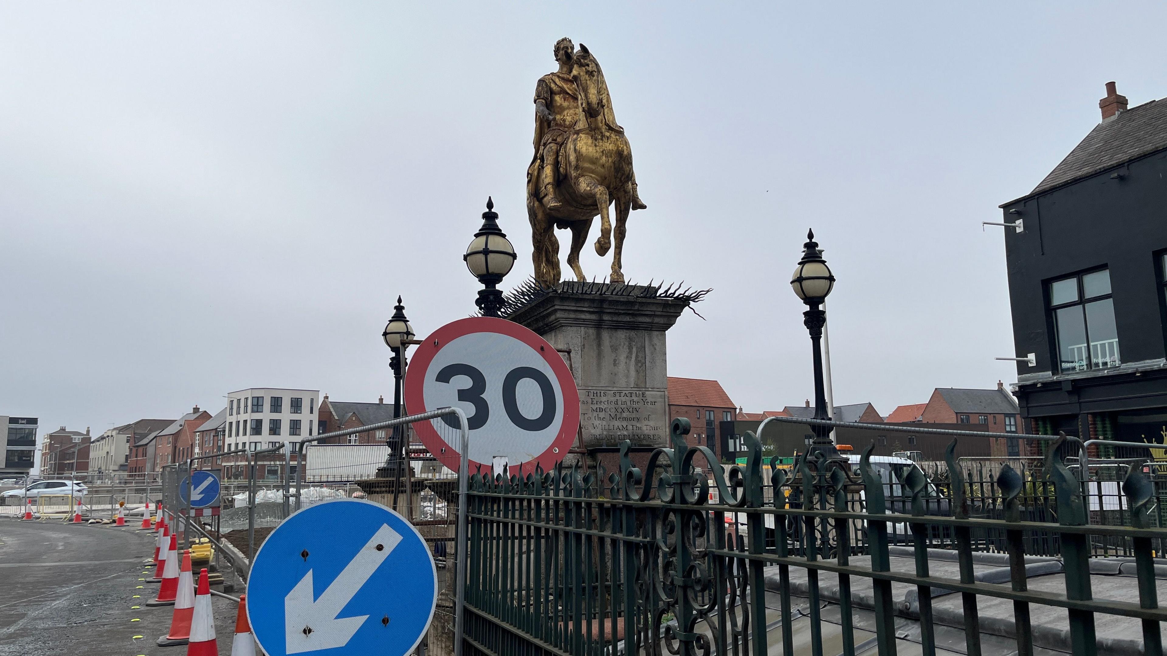 The statue of King William III sits amongst roadworks and is surrounded by orange traffic cones and temporary signs. King William is sat on a horse and the colour of the statue is gold