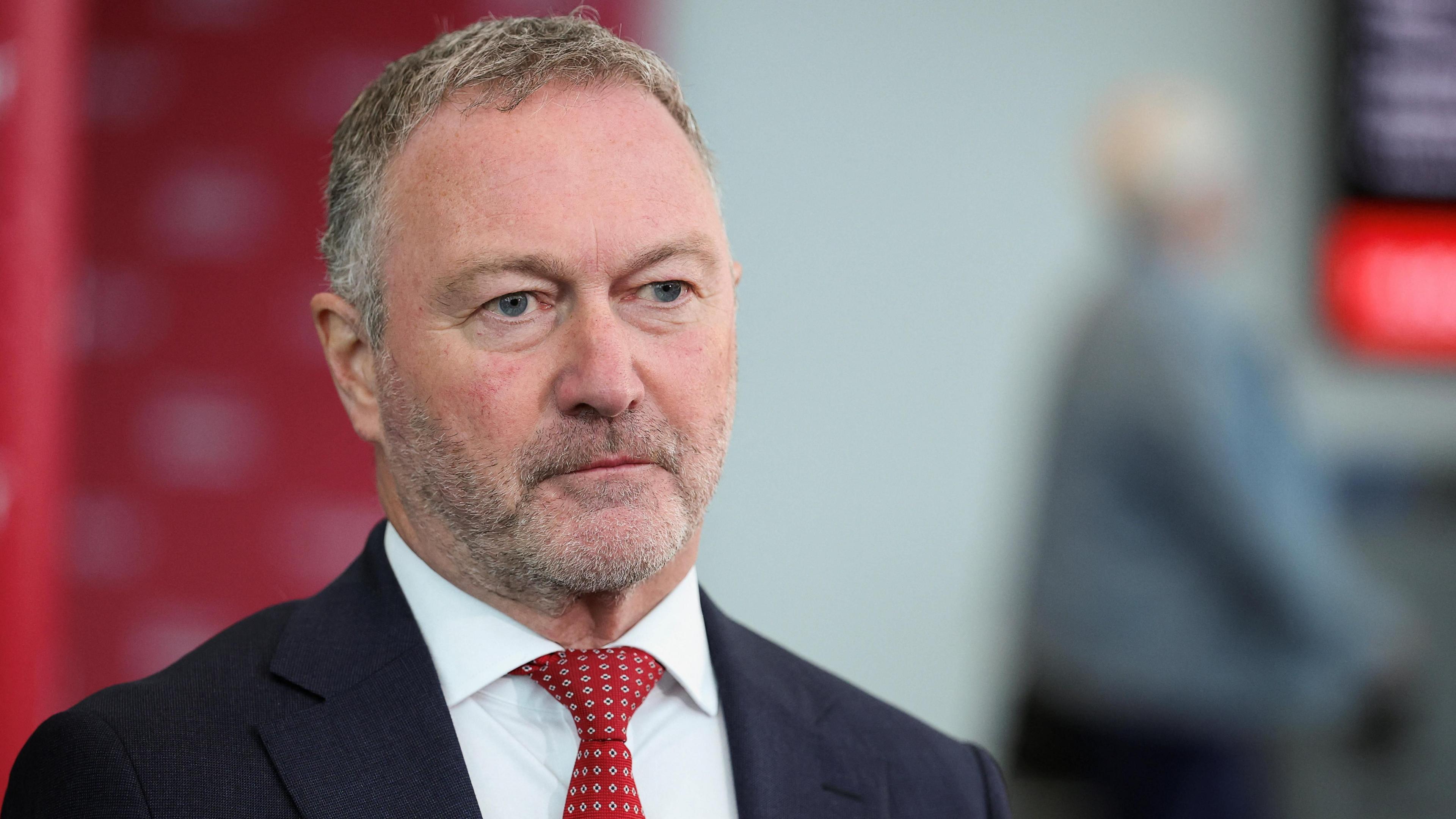 Steve Reed in a white shirt and red tie. He is unshaven with short light brown hair. He is standing inside, looking slightly off camera.
