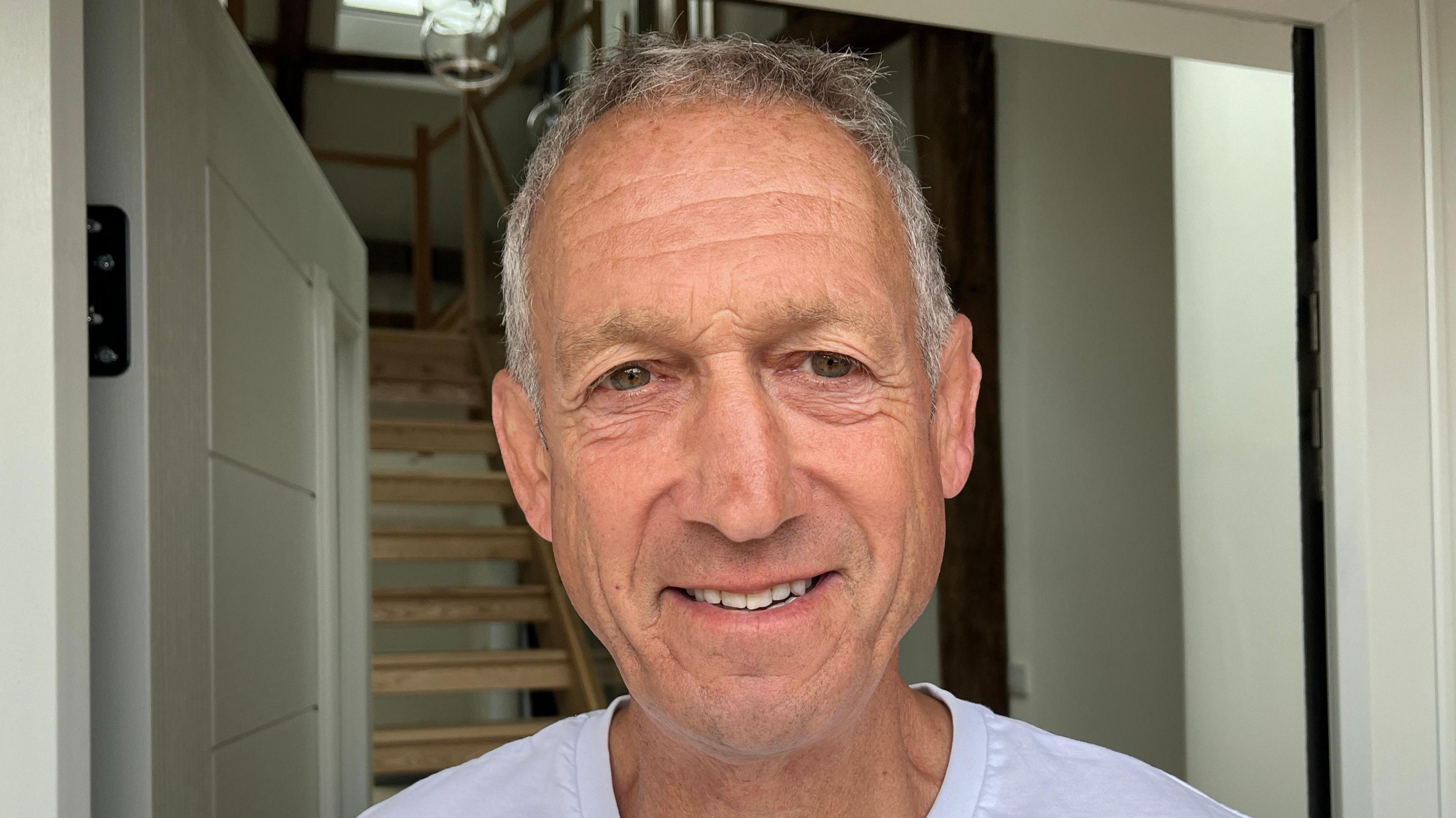 Harry Bowden smiles at the camera in the doorway of the barn conversion. He has grey hair and wears a white T-shirt.