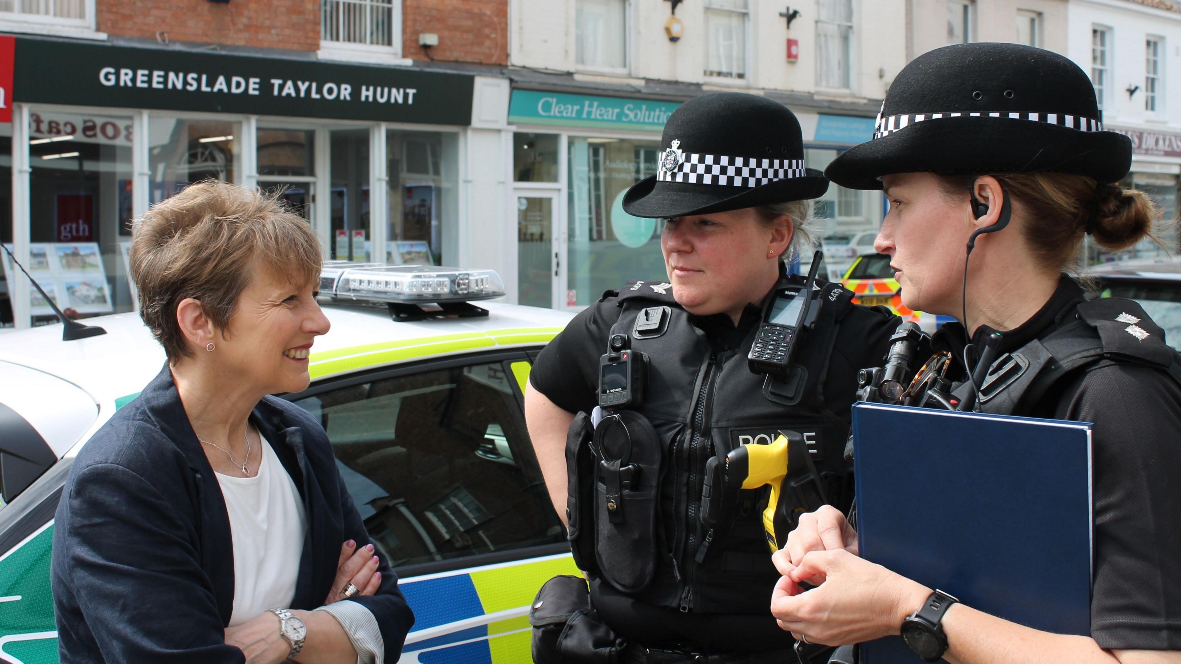 Clare Moody, wearing a black blazer over a white top, stands smiling with her arms folded on the left of the image as she talks with two uniformed female police officers. Shops can be seen in the background.