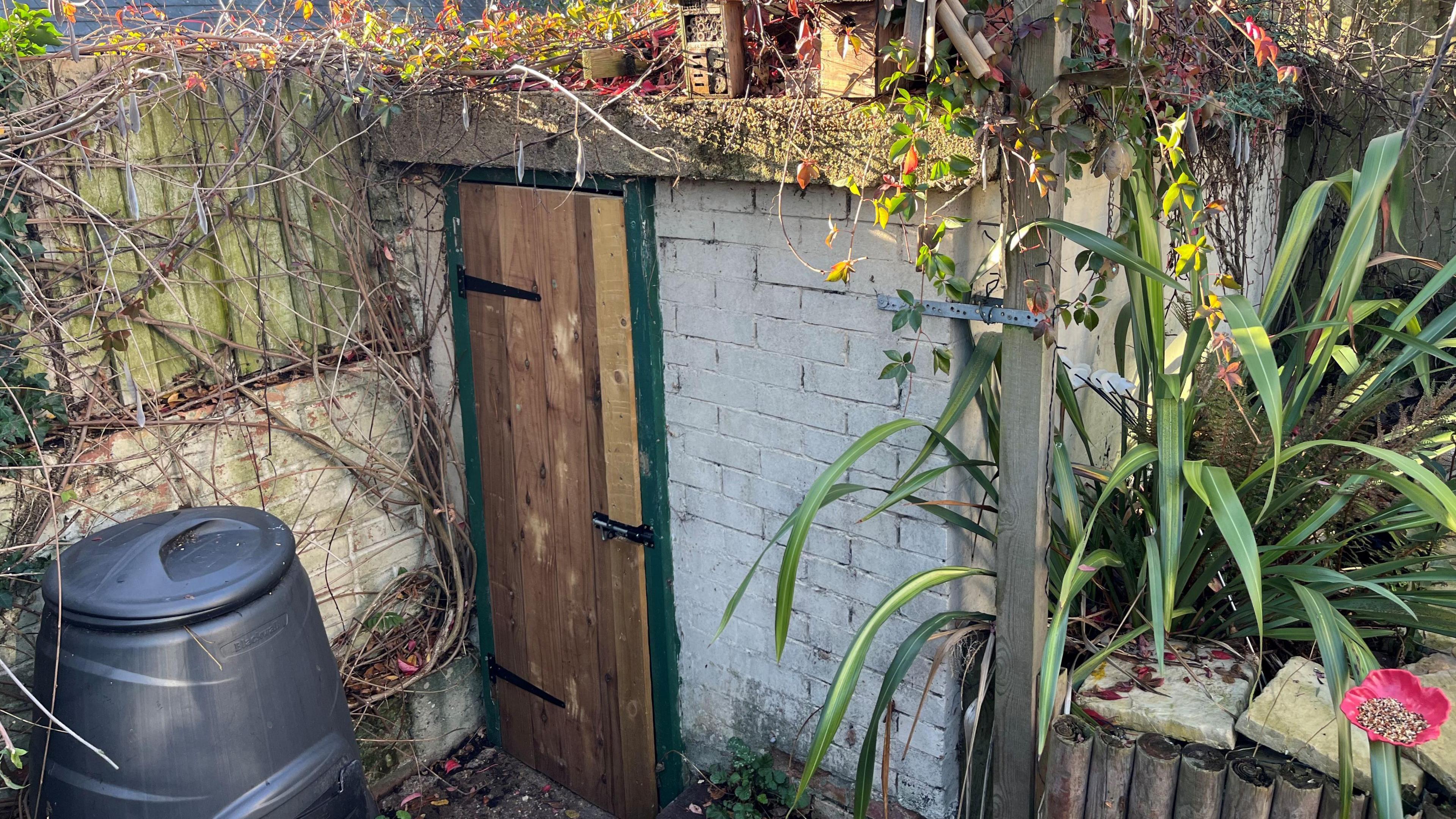 A white washed brick building about 2m (6ft) by 2.44m (8ft), it has a green doorframe and a new wooden door with a metal bolt. It has a 22cm (9in) thick reinforced concrete roof. There is a black compost bin on the left and some shrubs on the right.