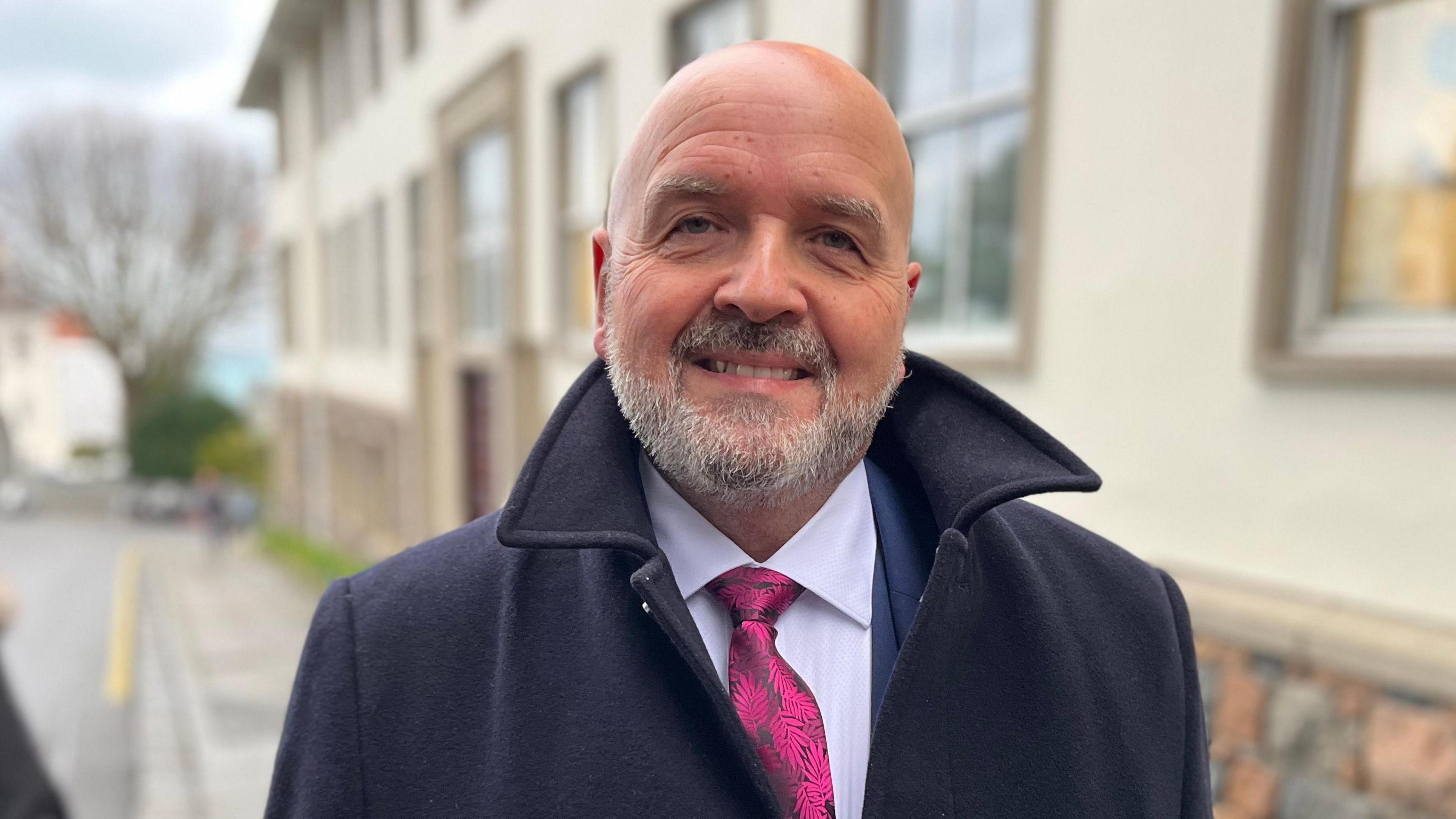 A bald man who is smiling, with a grey beard. He's wearing a blue jacket with a high collar and a white shirt with a metallic pink tie with a flower pattern. 