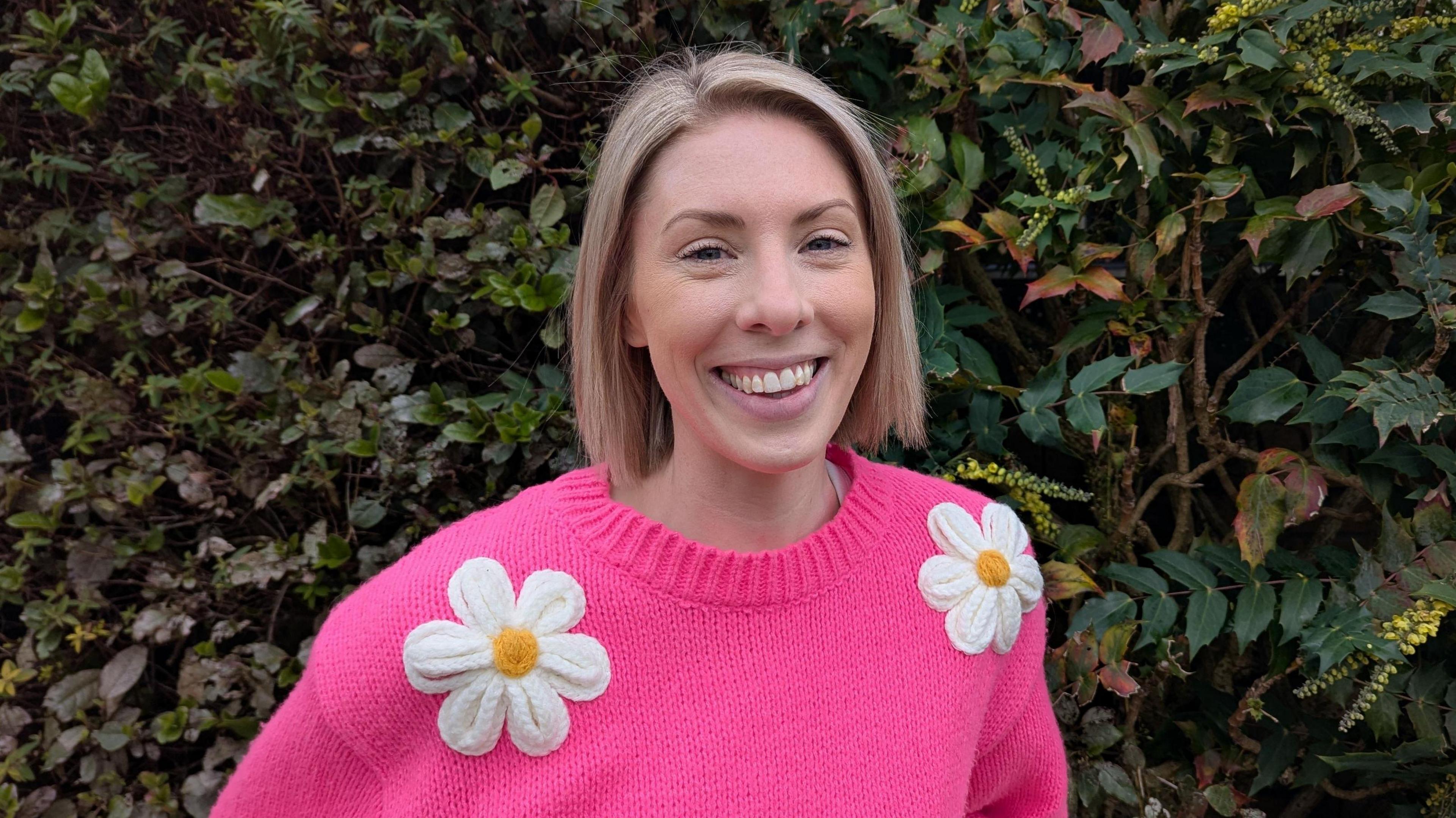 Daisy, pictured on her own in front of a hedge. She is wearing a bright pink jumper with large daisies on it and is smiling at the camera. She has short blonde hair. 