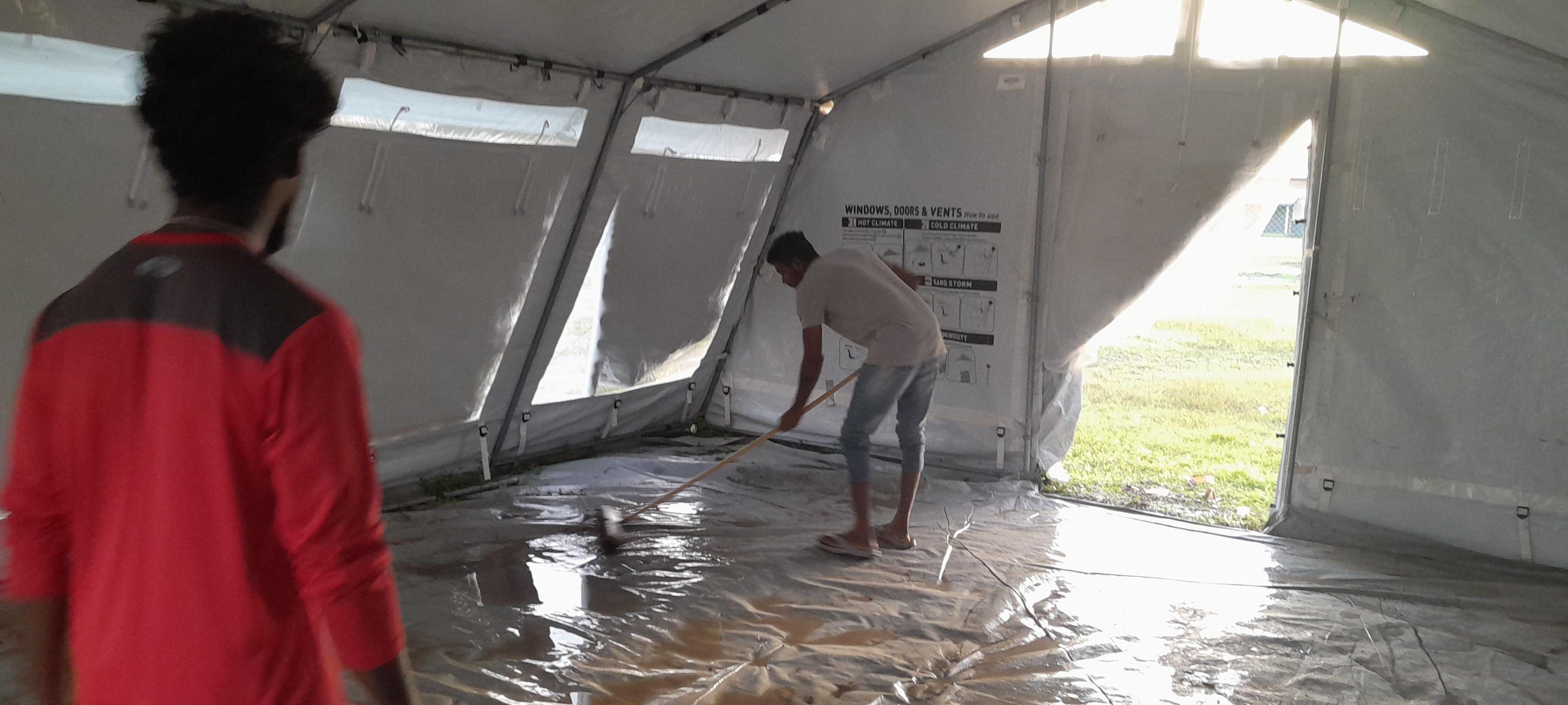 Two men are stood inside a large tent. One of them is using a brush to sweep large puddles of muddy water