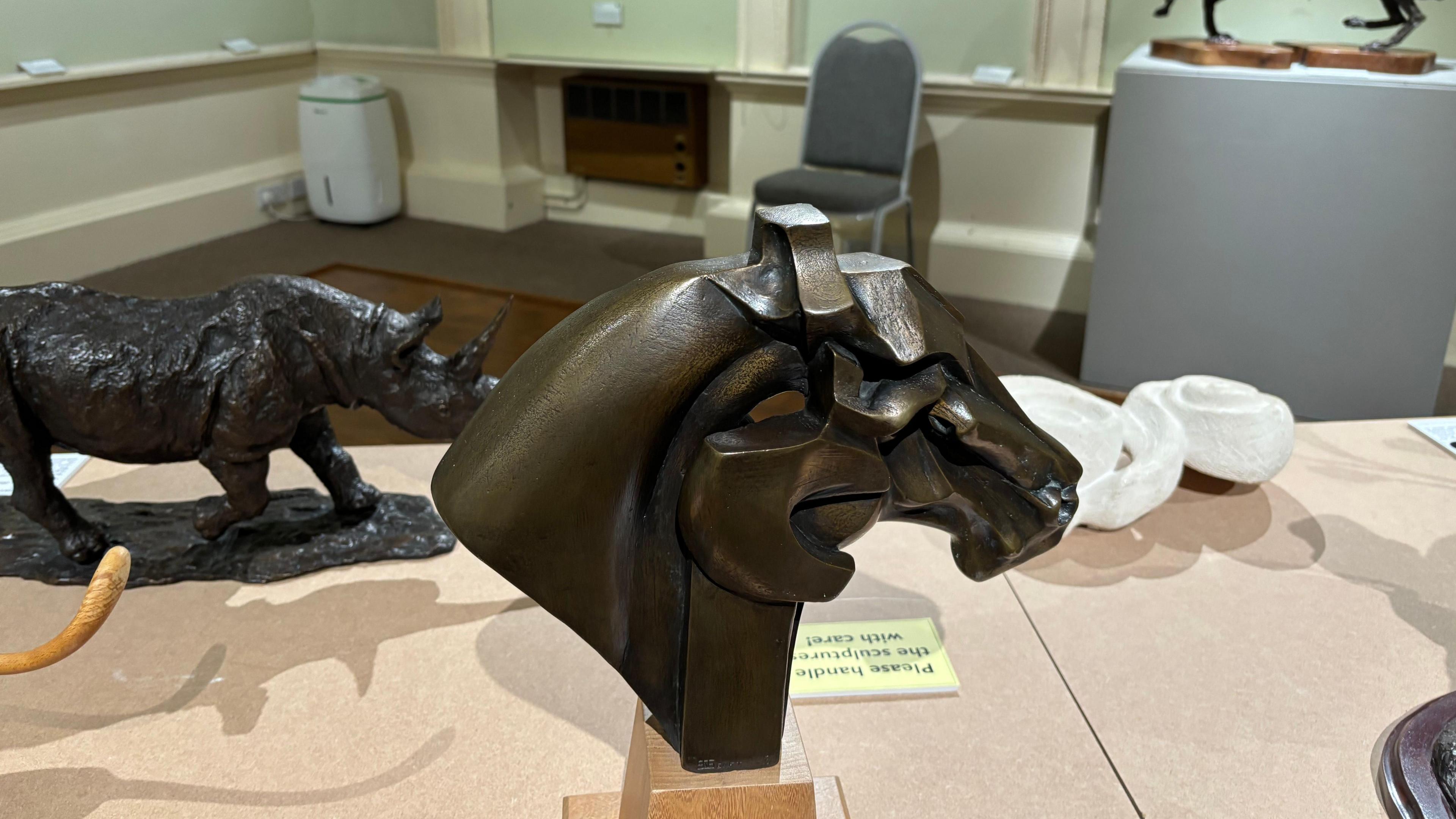 The image depicts a refined art gallery space showcasing a mix of sculptures and paintings, with a bronze sculpture of two abstractly merged horse heads prominently displayed in the foreground atop a wooden base, accompanied by a sign urging visitors to handle exhibits with care. Behind it, a realistic rhinoceros sculpture strides forward, while to the right, two dynamic horse figures rear up in an expressive interaction. The backdrop features framed artworks, a grey chair, and a white appliance—likely a dehumidifier or air purifier—set against classic wall moulding and a carpeted floor, creating an atmosphere of quiet elegance and thoughtful curation.