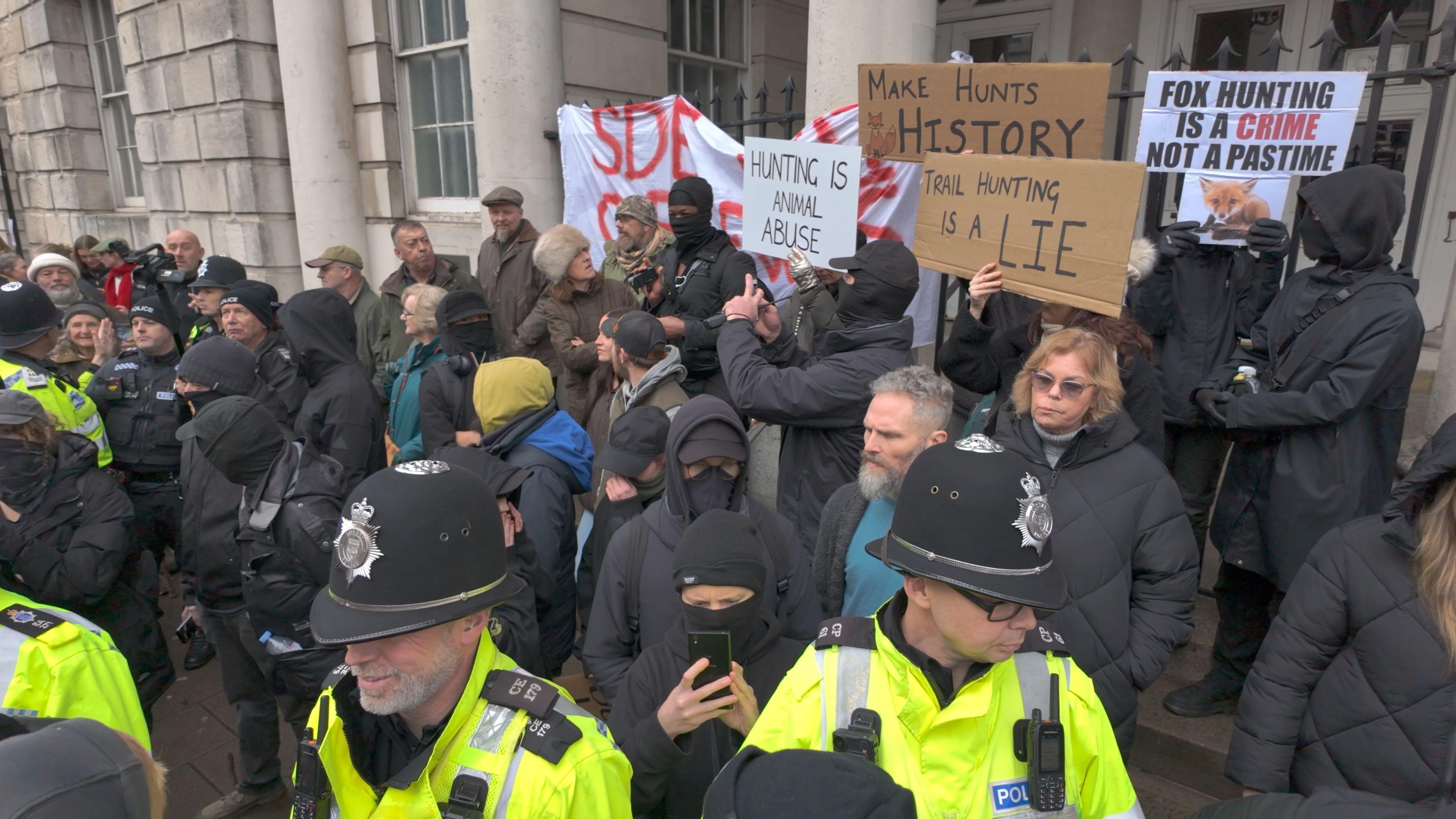 Police stand in front whilst people dressed in black with anti-hunt placards stand behind on steps.