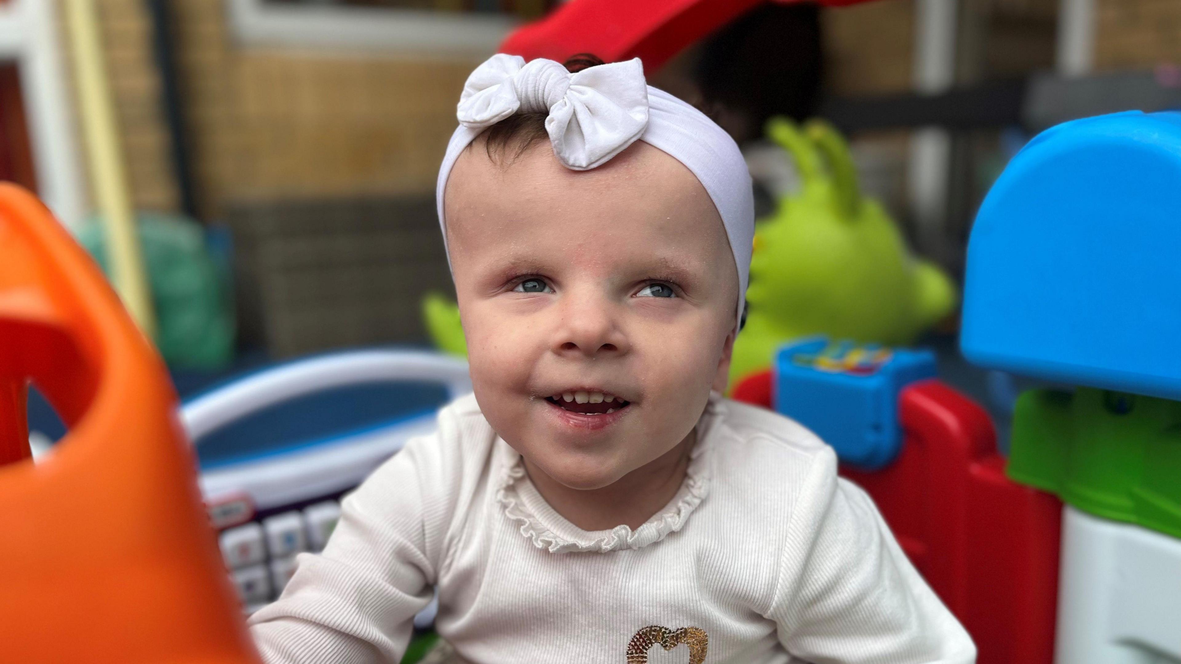 Four year old Olivia smiles at the camera. She is wearing a white top with a gold heart on the right hand side, and has a white bow on her head. Although the background is blurred, you can see she is sat inside a colourful playhouse.