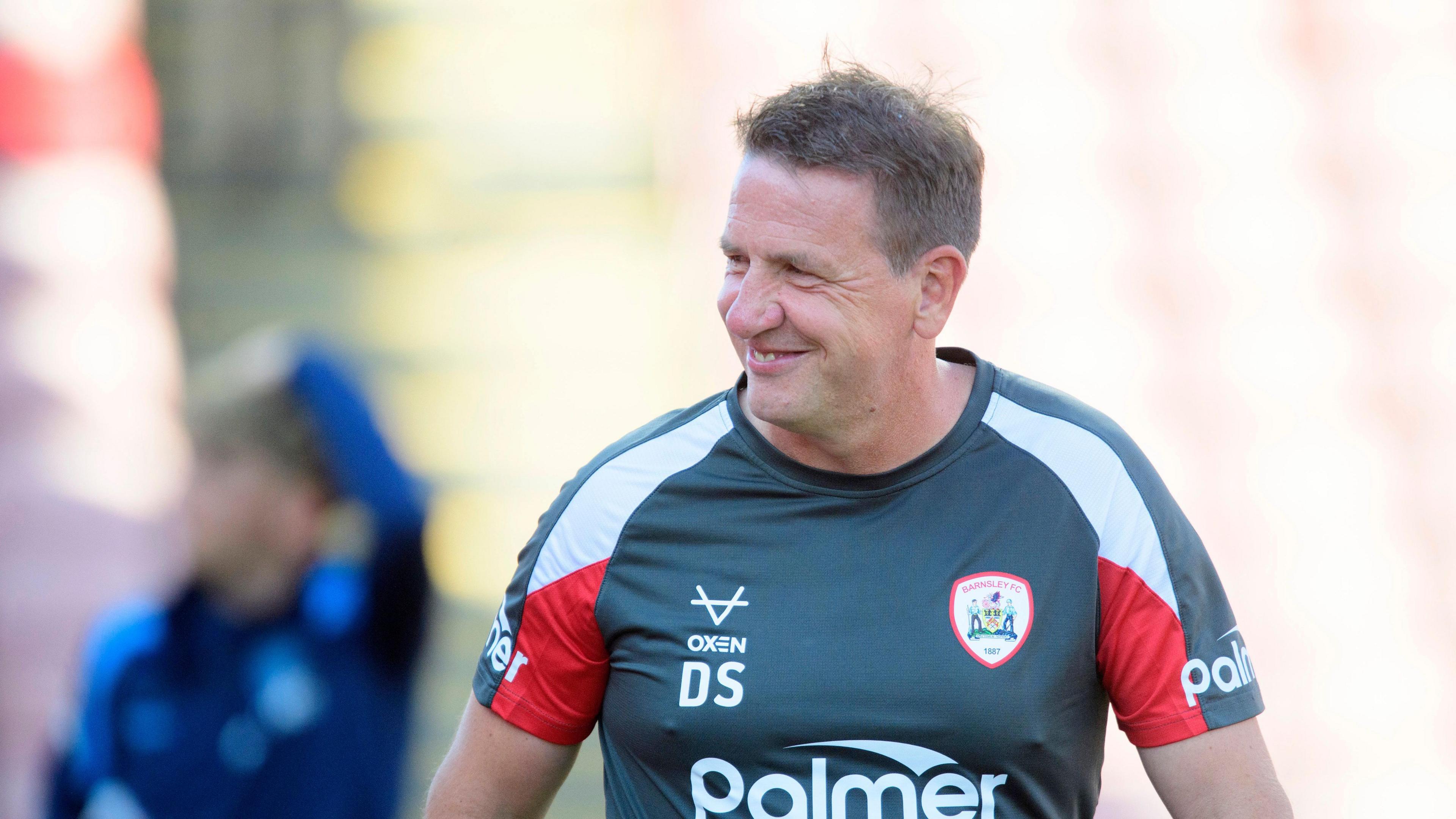 Daniel Stendel smiling as he looks away from the camera while wearing grey top with a Barnsley crest