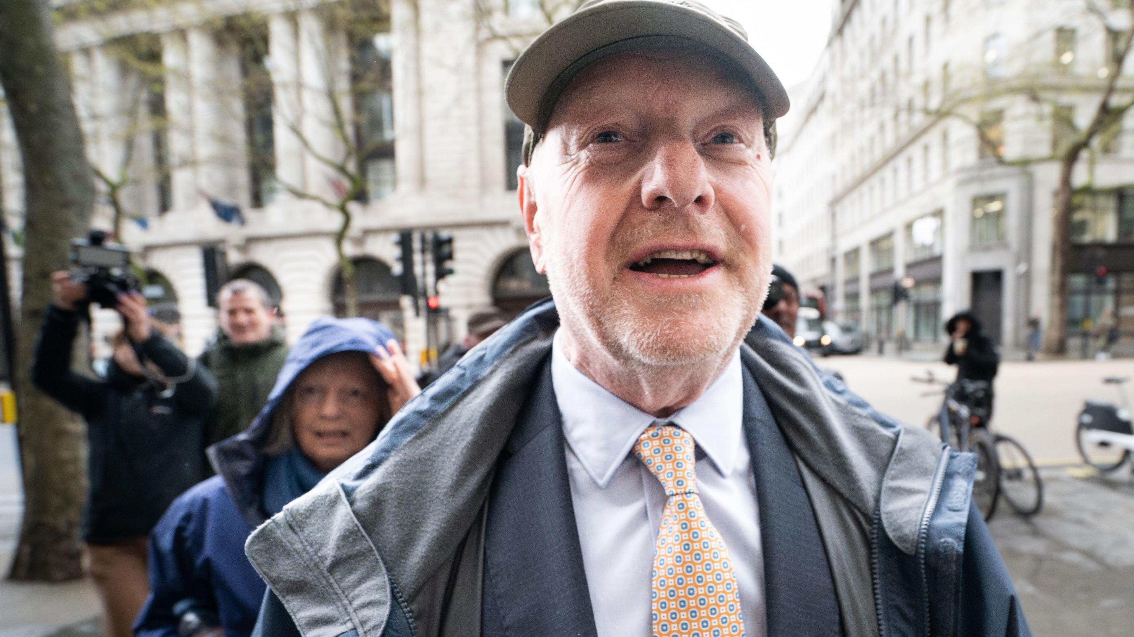 Former subpostmaster and lead campaigner Alan Bates, wearing a suit and tie, arrives at Aldwych House, central London, to give evidence to Post Office Horizon IT inquiry. He has stubble on his chin and a baseball cap on his head while behind him a woman shelters under a hood on her coat.