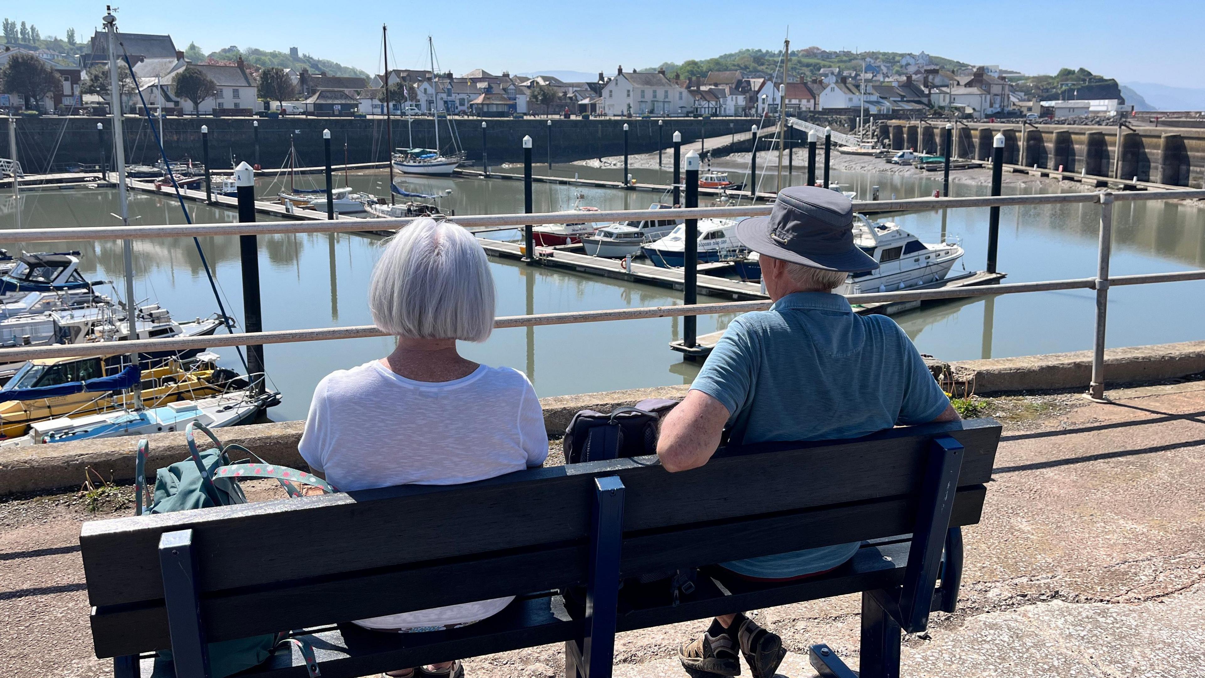 A photograph of a couple sitting on a bench looking over a small harbour on a sunny day. The photograph is taken from behind.