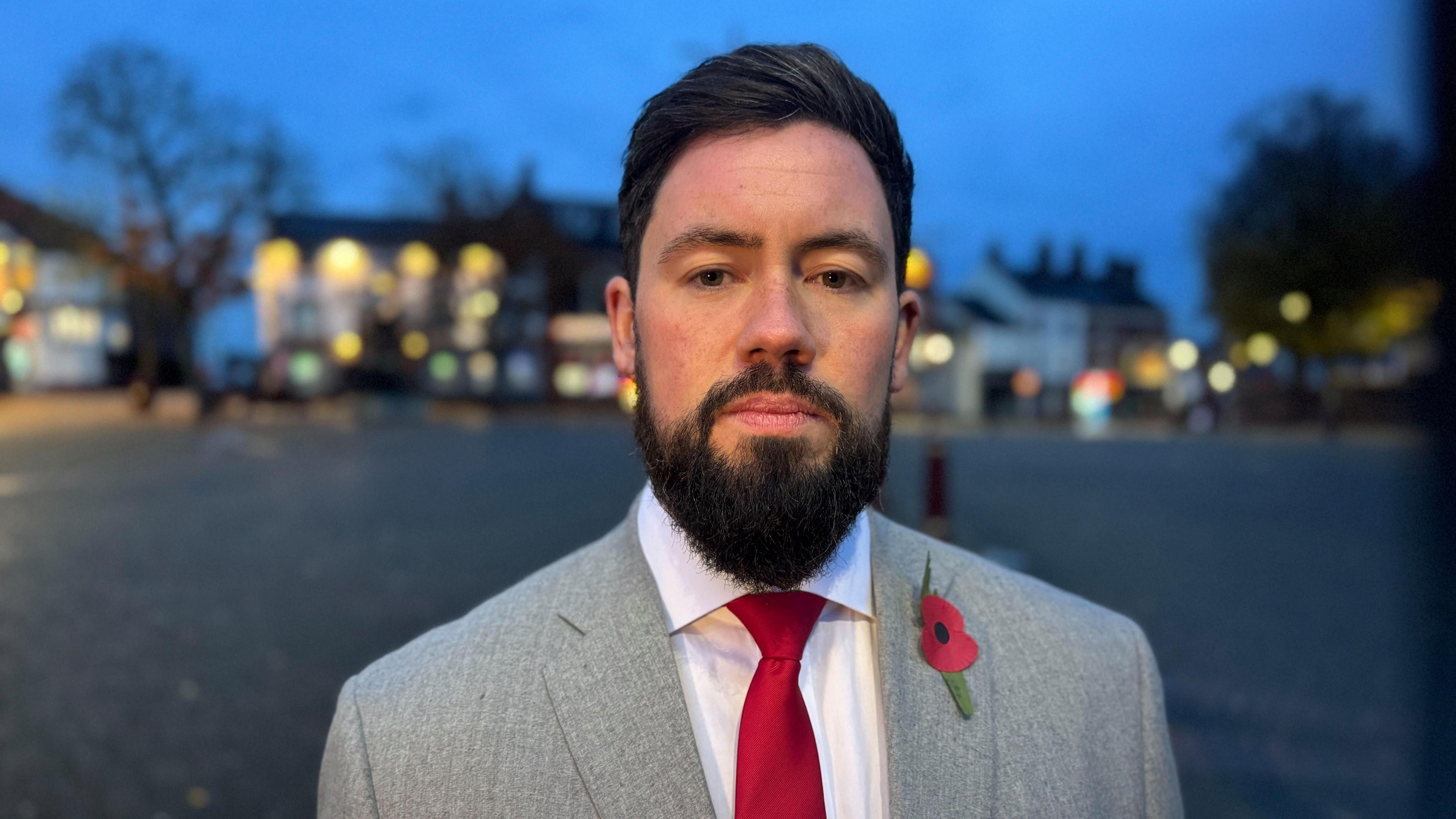 Adam Thompson, MP wearing a grey jacket, white shirt and red tie and sporting a poppy stands in Ilkeston Market place just as night falls. The lights are on in the pubs and bars behind him.