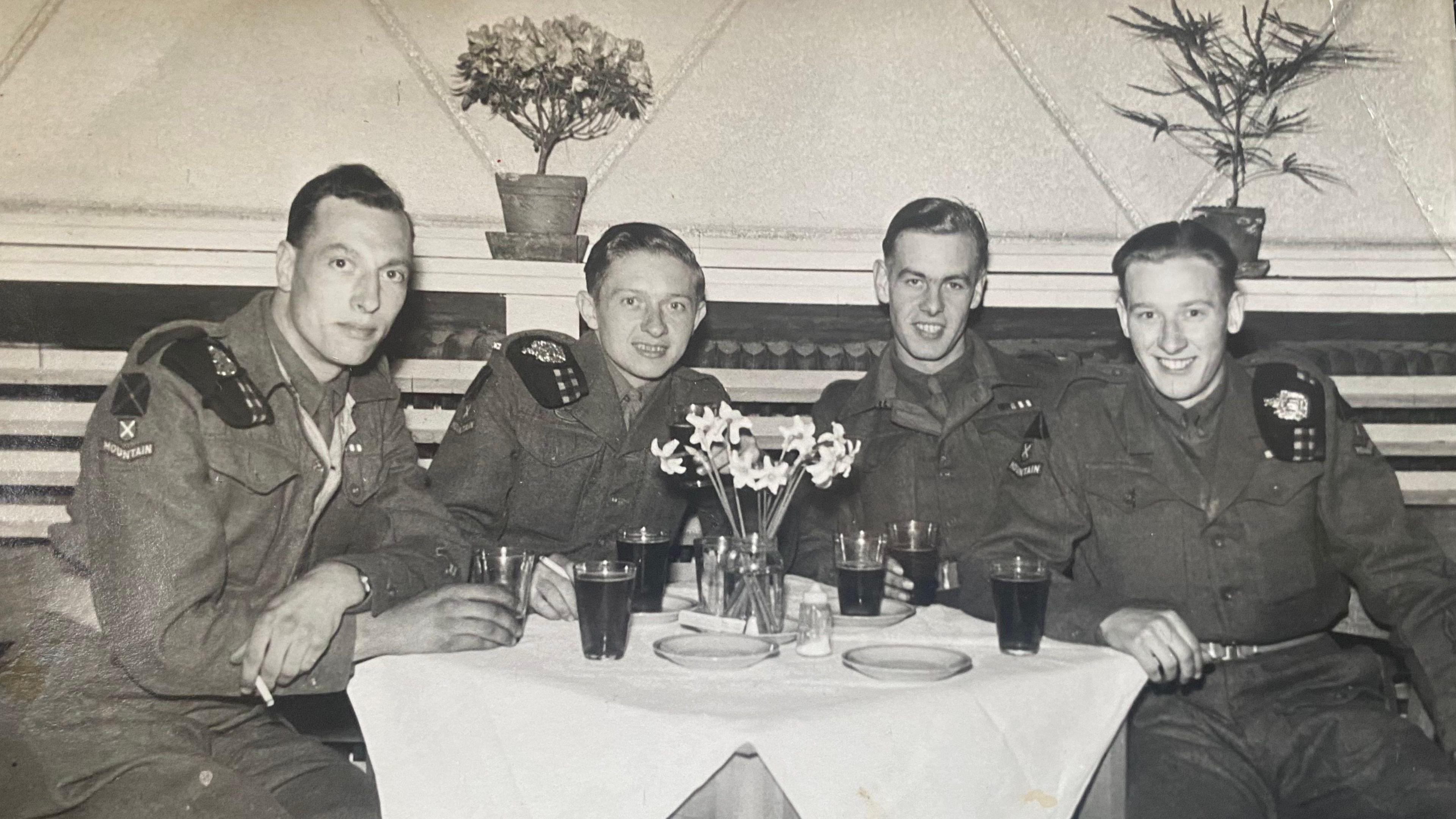 A black and white photo of 4 young men in solider uniforms sat around a table with drinks. There is a vase of flowers in the middle of the table.