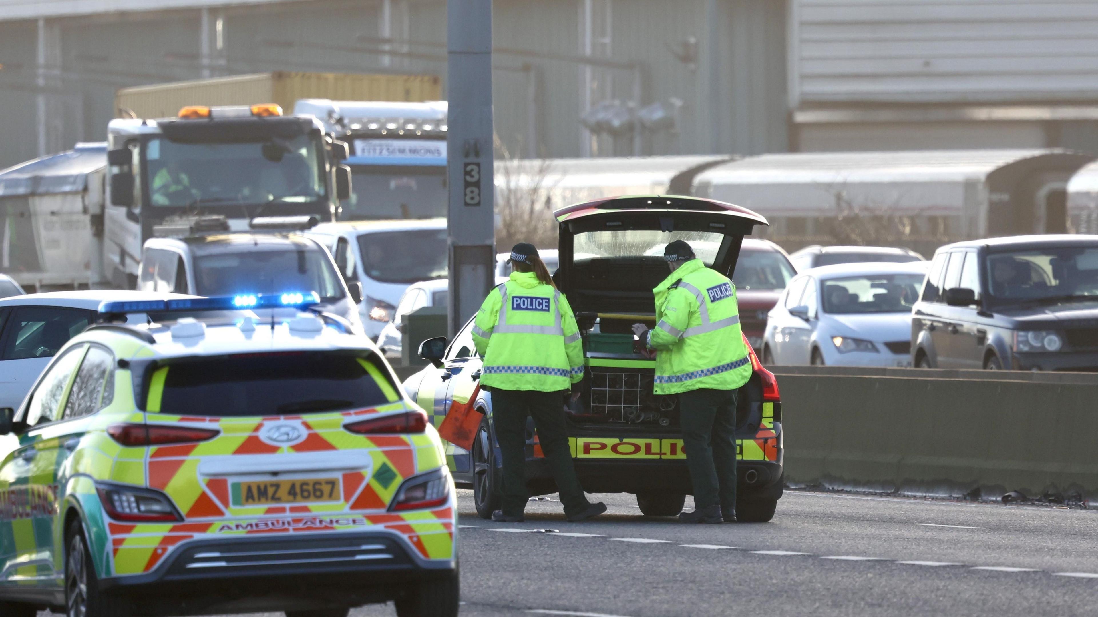 An ambulance car and a police car sitting on the tarmac of the M2 - two police officers in high-viz jackets stand with their backs to the camera at the open boot of the police car. Behind them queuing traffic can be seen.