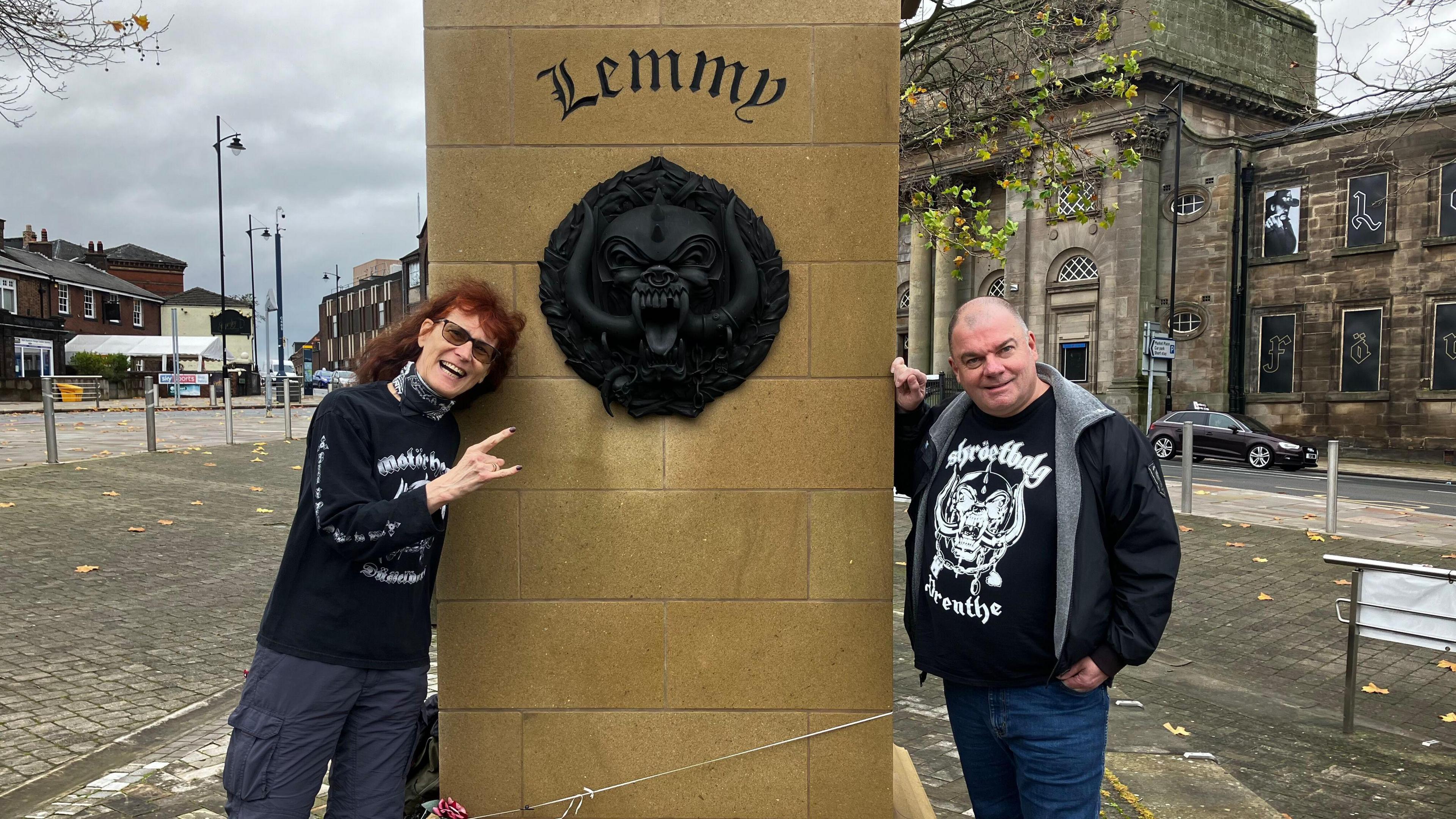 A couple in Motorhead themed T-shirts stand either side of the plinth of the Lemmy statue, which has Lemmy engraved on it. Also on the plinth is is Motorhead's large black motif, which is known as the Snaggletooth or the War Pig and sometimes the Iron Boar. The statue is in a square, with roads and buildings surrounding it.