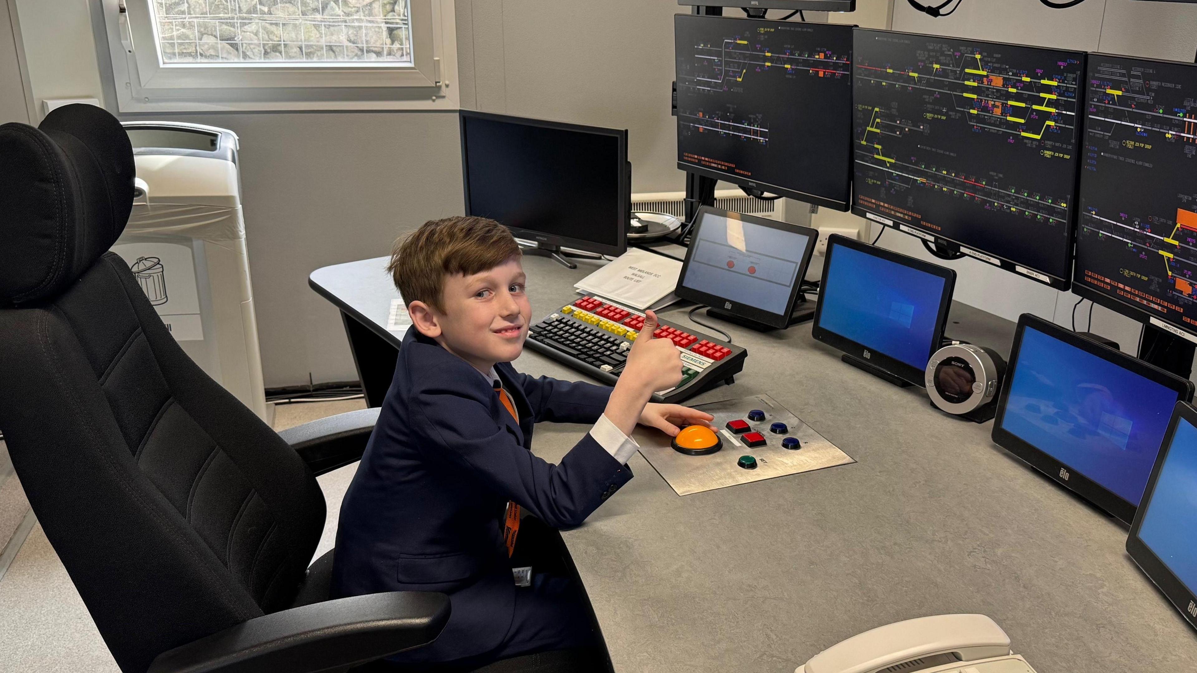 A young boy with brown hair in a blue suit sat in front of a large desk with a number of large computer monitors in front of him