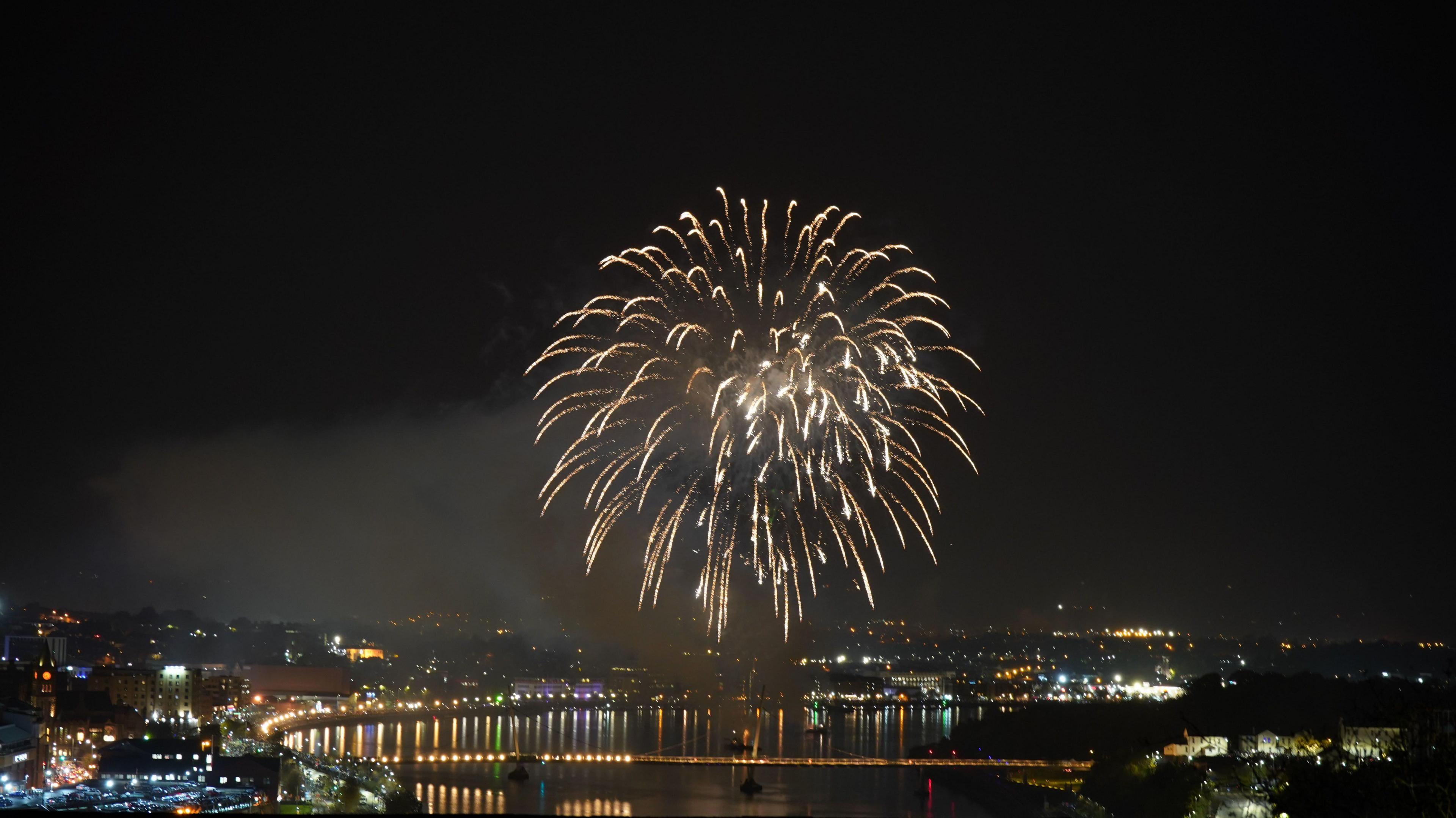 A singular large firework explodes over the Foyle Bridge in Londonderry. The city is in darkness bar the lights of the bridge and buildings, which are reflected in the River Royle.