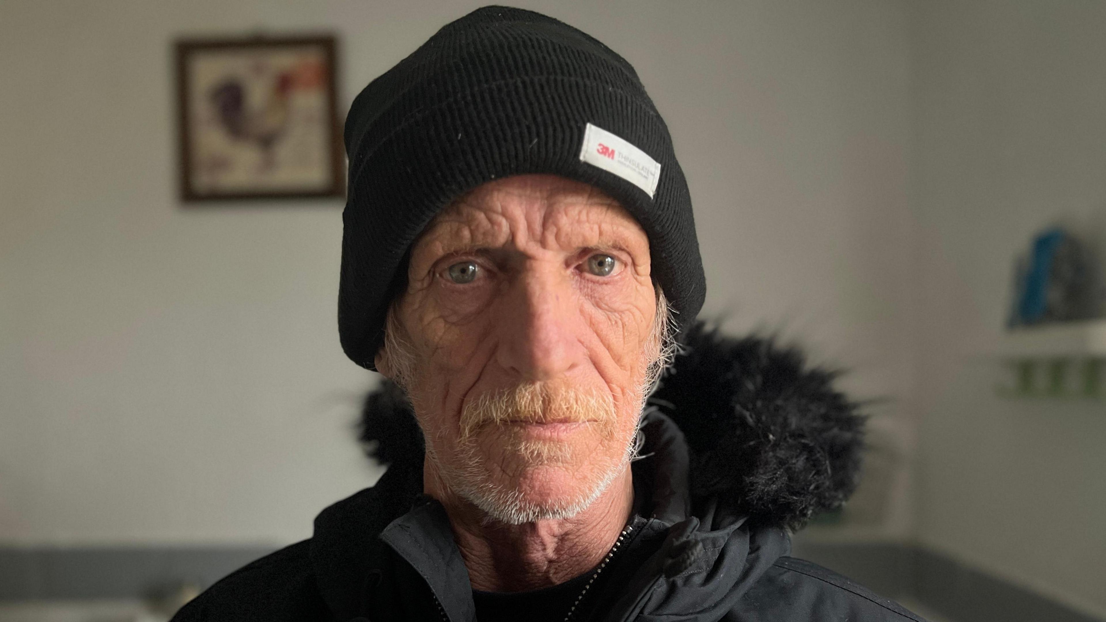 Ray is looking at the camera, he is an older man and is wearing a black woolen hat and a black coat. He has a short white beard. He is sitting inside in a room with white walls and a picture on the wall.