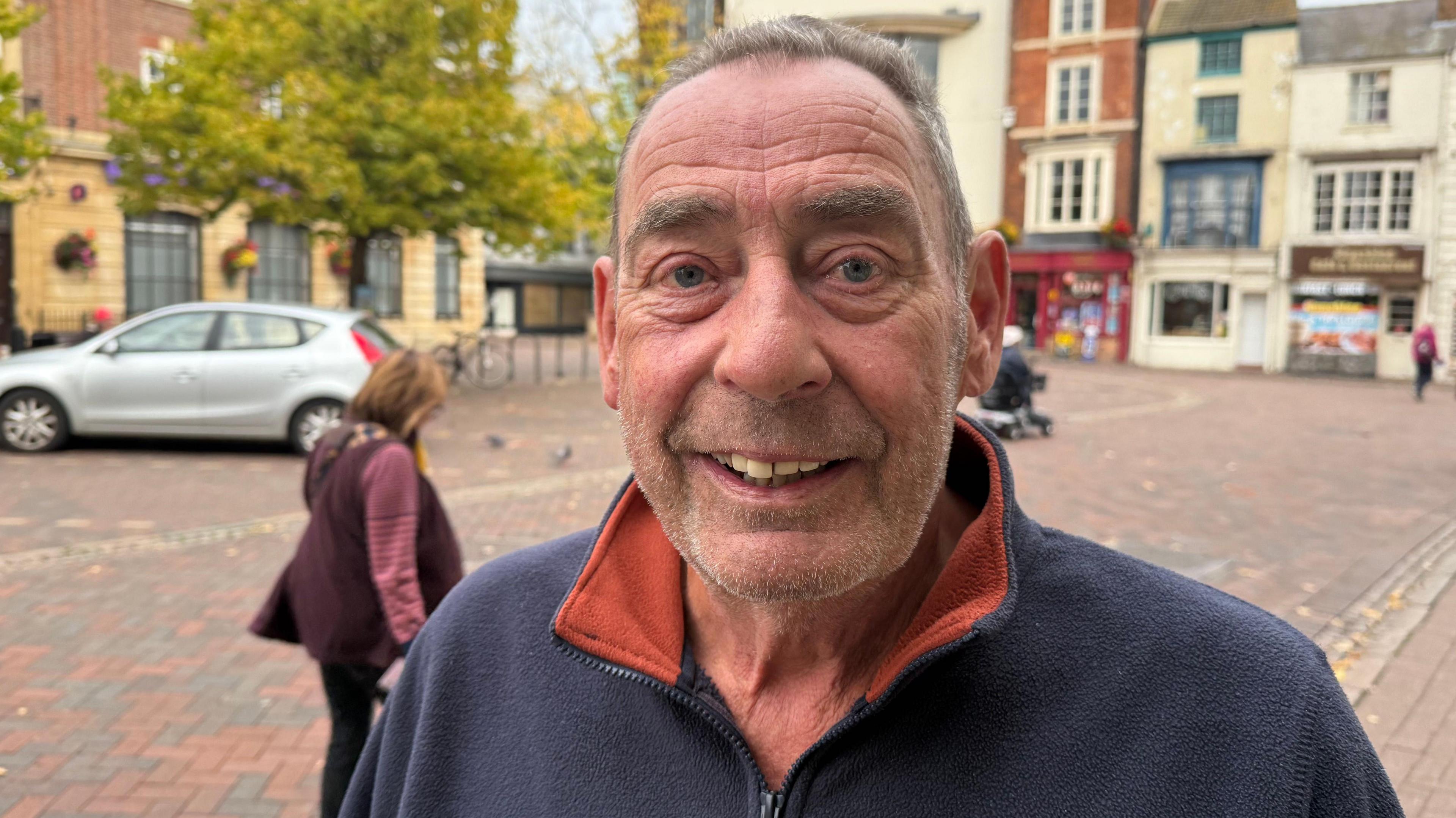 A man with short brown hair and stubble smiles broadly as he stands in a town square. He is wearing a blue zip top. A silver car is parked in the background and pedestrians are walking past.