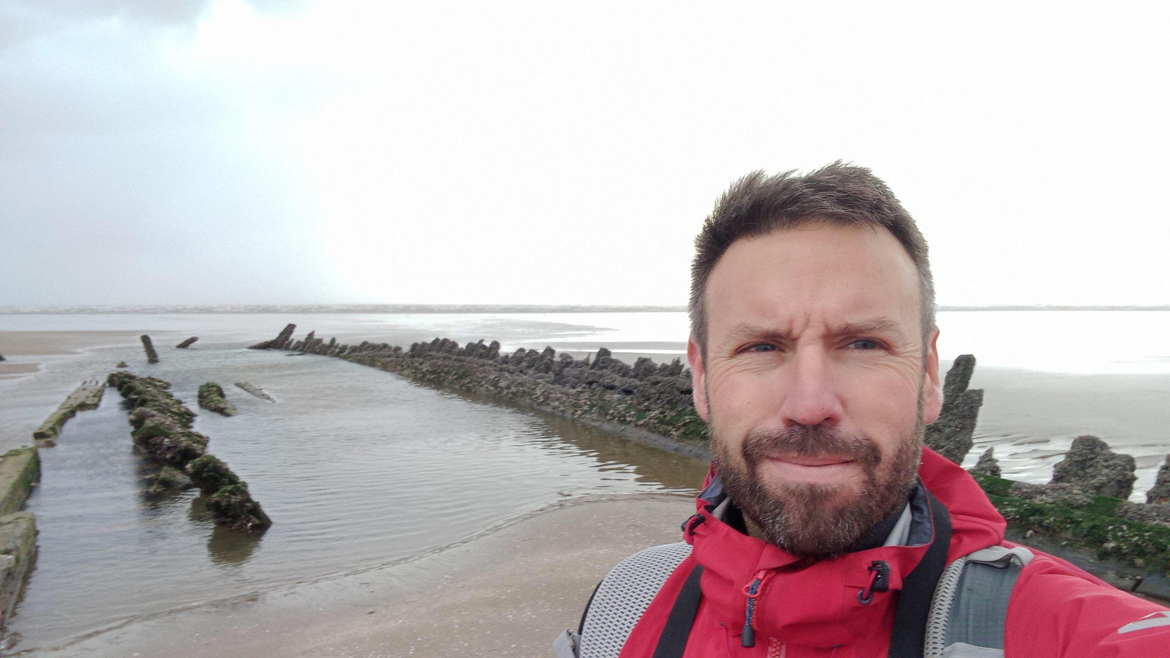 Dr Julian Whitewright smiling at the camera (selfie style) in front of Cefn Sands shipwreck which is visible poking out of the sand, pools of water within the wreck show where sand has been hollowed out.