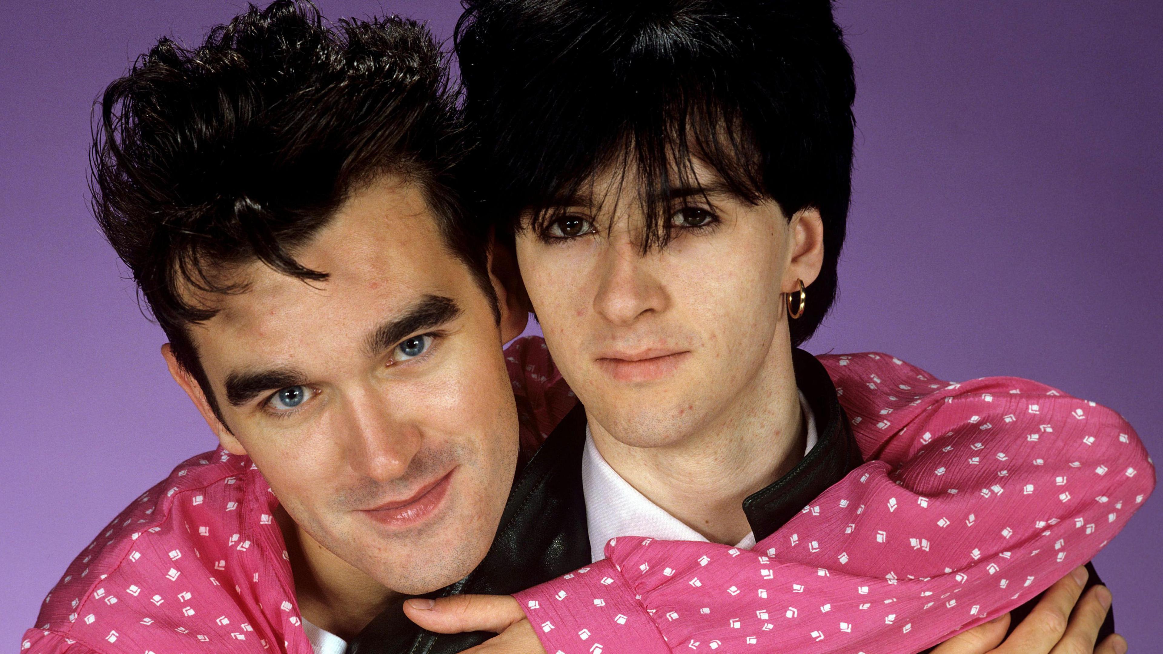 Morrissey, with dark quiffed hair and a pink and white shirt, hugs Johnny Marr, who has black hair with a fringe across his eyes, a gold earring and a white T-shirt underneath a black leather jacket