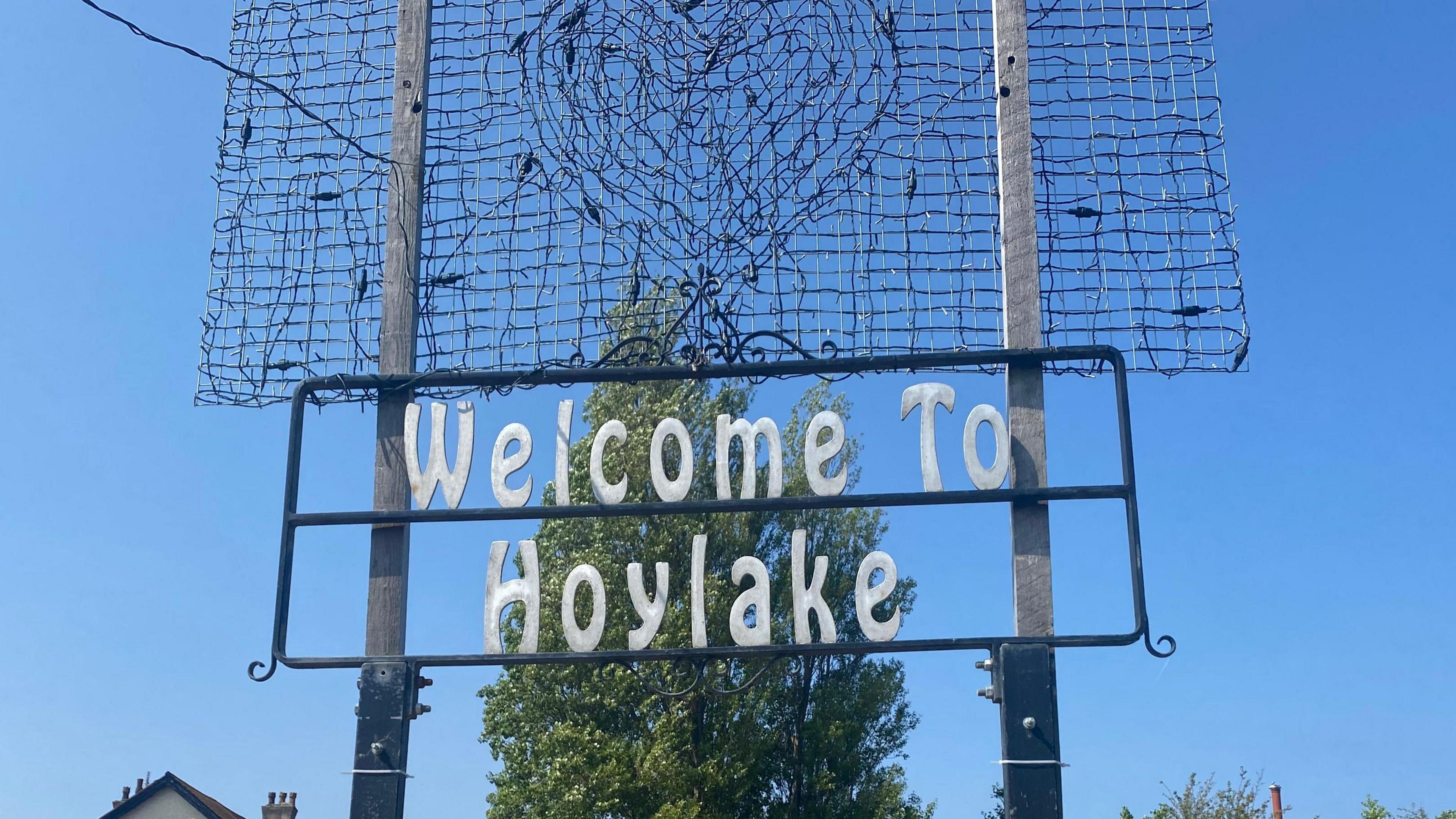 A white-lettered sign reading 'Welcome to Hoylake' hangs between two grey posts, underneath an unlit heart-shaped light installation and in front of a green-leafed tree and a cloudless blue sky